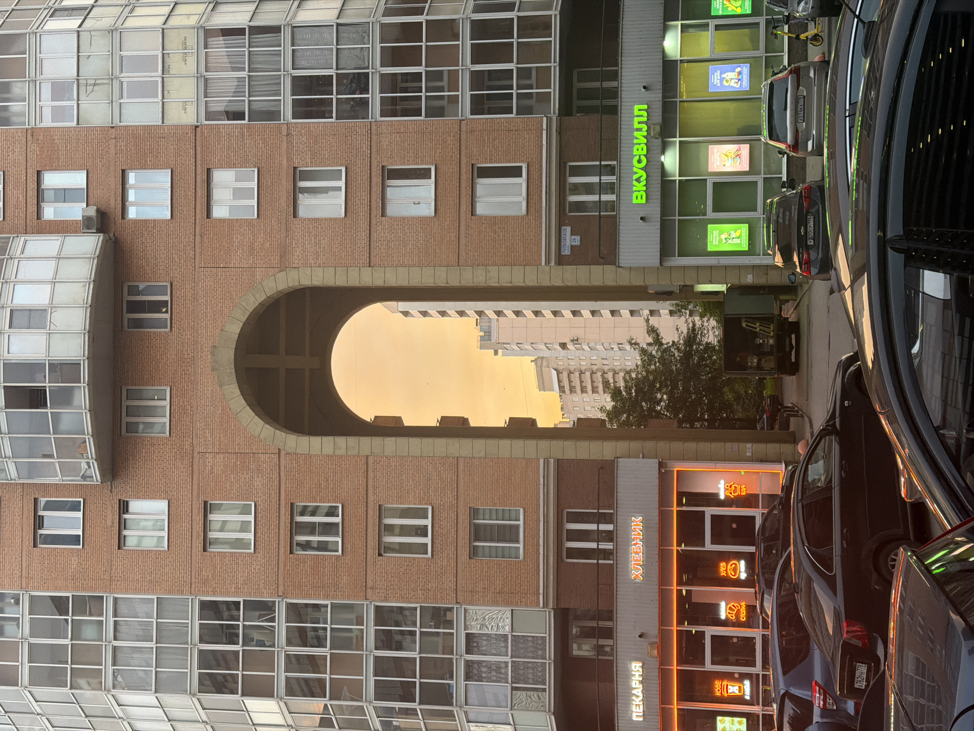 The image shows an exterior view of a brick apartment building with a large archway in the center. Through the archway, a light orange sky is visible, hinting at either sunrise or sunset. On the ground level beneath the archway, a street scene unfolds. To the left, a storefront with neon signs reads "ПЕКАРНЯ" (Bakery) and "ХЛЕБНИК" (Bread Maker). The storefront has large glass windows displaying neon signs with images of coffee, bread, and food. To the right of the archway, another storefront with a green illuminated sign reads "ВКУСВИЛЛ". This store also has large windows displaying posters and advertisements. Several cars are parked in front of the stores, including a black sedan, a silver SUV, and a dark-colored sedan in the foreground. A delivery truck is parked near the right side of the archway. In the background, taller residential buildings are visible. The overall scene depicts a street in a city, likely in a residential area, with commercial establishments on the ground floor of an apartment building.