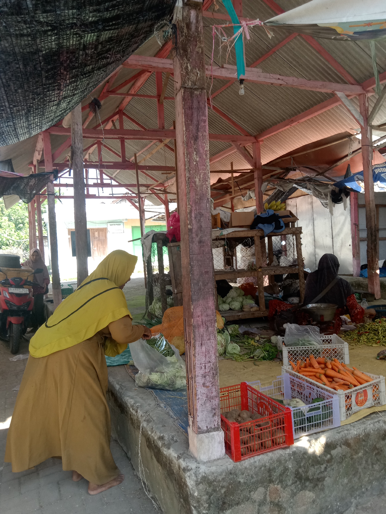In the city of Klepek, Indonesia, a market stall is depicted under a rustic, open-air shelter. A woman wearing a yellow hijab and a long, mustard-colored garment is seen from the back, facing towards the right of the frame. She is interacting with produce, appearing to be bagging items from a large orange bag. In front of her, on a concrete ledge, are several crates of fresh vegetables. A red crate contains potatoes, while a white crate holds carrots. To the left, a red motorcycle is parked. Further back in the market, another woman wearing a dark hijab is seated, with various vegetables displayed around her. The overall environment suggests a bustling, local marketplace with a focus on fresh produce.