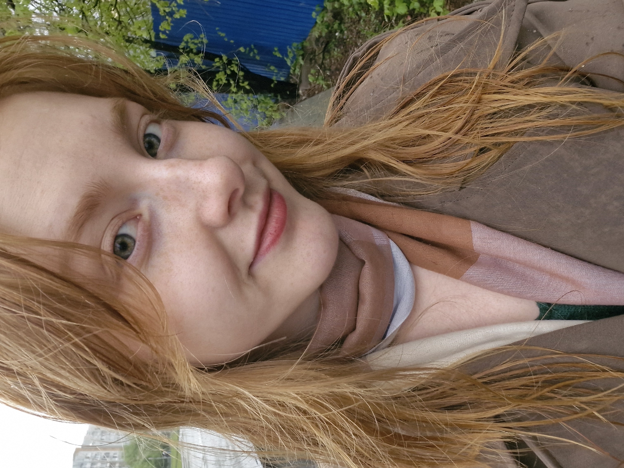 A close-up portrait of a young woman with fair skin, freckles, and long reddish-blonde hair. She has light green eyes and is wearing lip color. She is wearing a brown jacket and a scarf with pink, brown, and white stripes. The background is blurred, but blue corrugated metal and green foliage suggest an outdoor setting. In the far distance, a building can be seen.
It is not possible to determine if the location is Yekaterinburg, Russia from the image alone. Further information would be required.