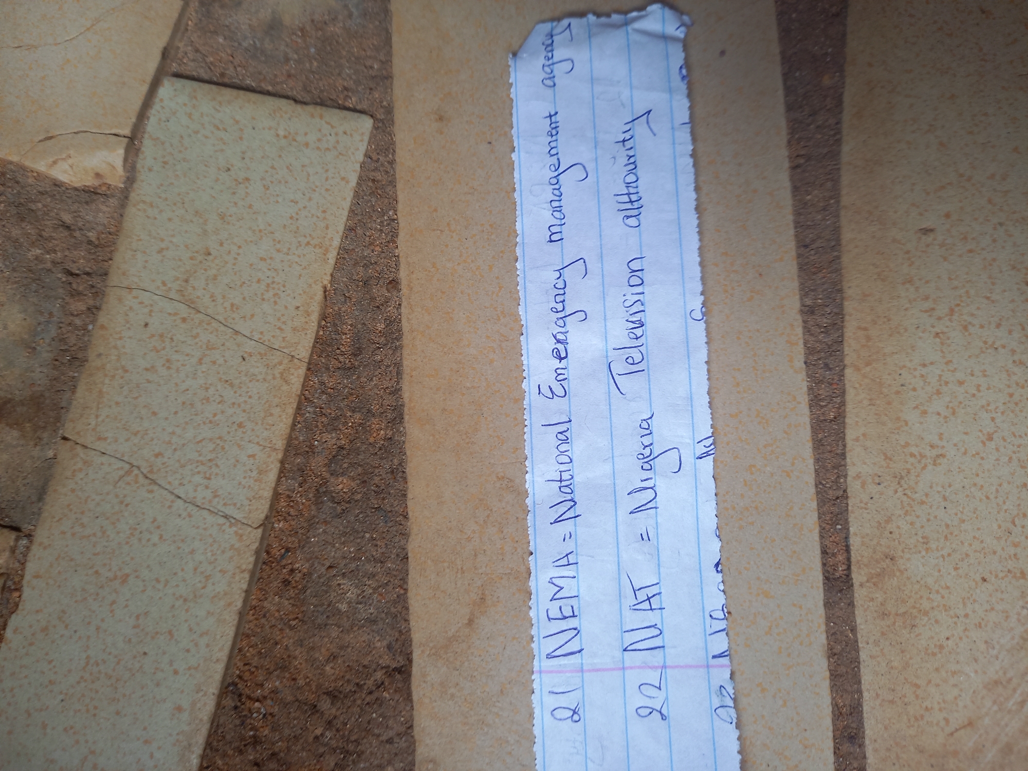 A torn piece of blue-lined paper, featuring handwritten text in blue ink, lies on a textured paved surface. The surface is composed of light-colored, speckled paving stones, possibly tiles, interspersed with darker grout lines or earth. The paper is somewhat crumpled and ragged at its edges, particularly the top right and bottom.
The visible text on the paper details acronyms and their full forms, numbered in a list format.
The clearly discernible entries are:
1. "21 NEMA = National Emergency management agency"
2. "22 NAT = Nigeria Television allhourity" (The last word appears to be "authority" but is written as "allhourity").
A third entry, "23 NCA," is partially visible at the bottom of the paper.
The setting appears to be an outdoor or semi-outdoor area, consistent with a path or ground covering common in places like Onitsha, Nigeria. The lighting is bright and even, suggesting it is daytime. There are no individuals or specific activities depicted beyond the implied creation of the notes on the paper. The scene offers a glimpse into a practical note-taking or study context within an everyday environment.