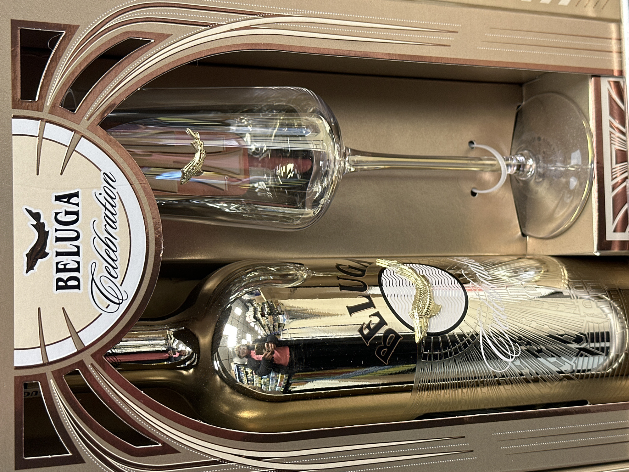 The image displays a Beluga Celebration gift set, likely of vodka, featuring a gold bottle and a glass. The set is presented in a decorative box with geometric designs. The scene is located in a retail setting, possibly in Moscow, Russia, given the Beluga brand's association with Russian vodka and the brand named on the package. A person holding a mobile phone is reflected in the surface of the shiny bottle.