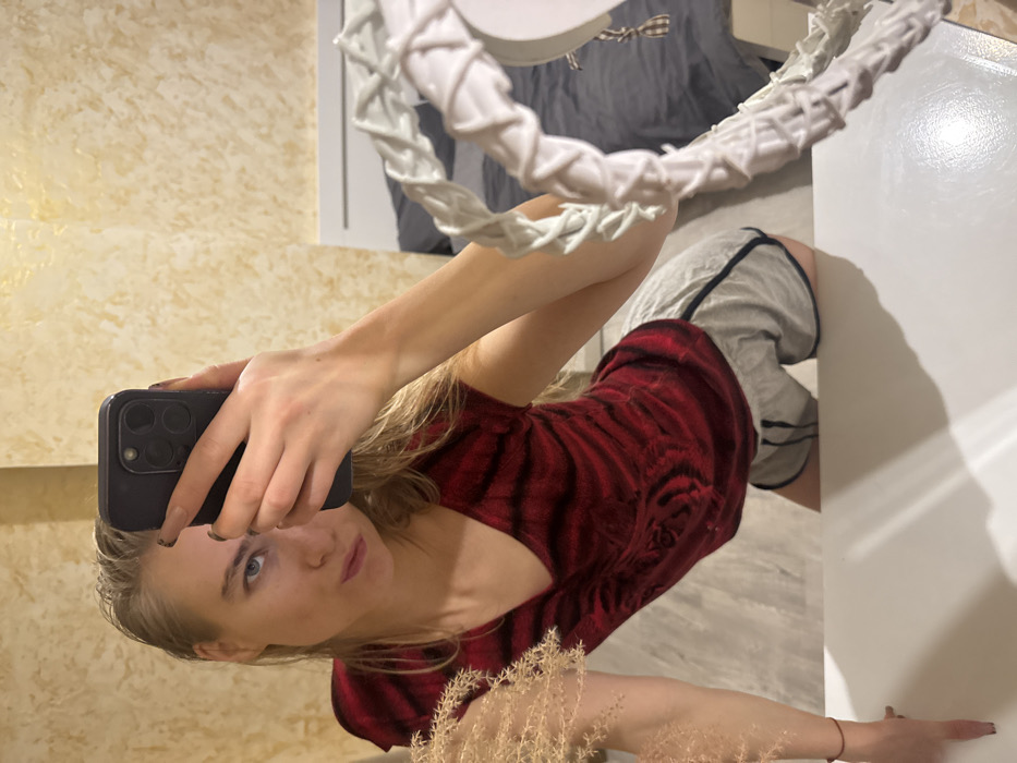 A woman holds a phone to take a selfie in front of a mirror. She has blonde hair and blue eyes, and she's wearing a red patterned top and gray shorts. Behind her, a wall is adorned with a textured beige design, and a white, ring-shaped decoration hangs from the ceiling. A portion of a white cabinet is visible on the right side of the frame. Some dried decorative plants are at the bottom of the photo.