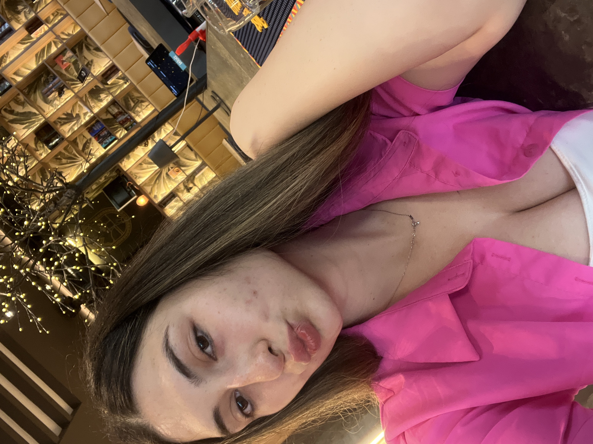 A young woman with long brown hair and fair skin is taking a selfie in a dimly lit indoor establishment. She is wearing a bright pink button-up shirt, partially unbuttoned to reveal a white top underneath. Her lips are pursed, and her skin shows some minor blemishes. The background is out of focus but appears to be a restaurant or bar, with bookshelves filled with items and decorative lighting. The image is an indoor photograph, taken from a close-up perspective, and does not contain any identifiable landmarks or elements that would allow for specific location determination within Plovdiv, Bulgaria.