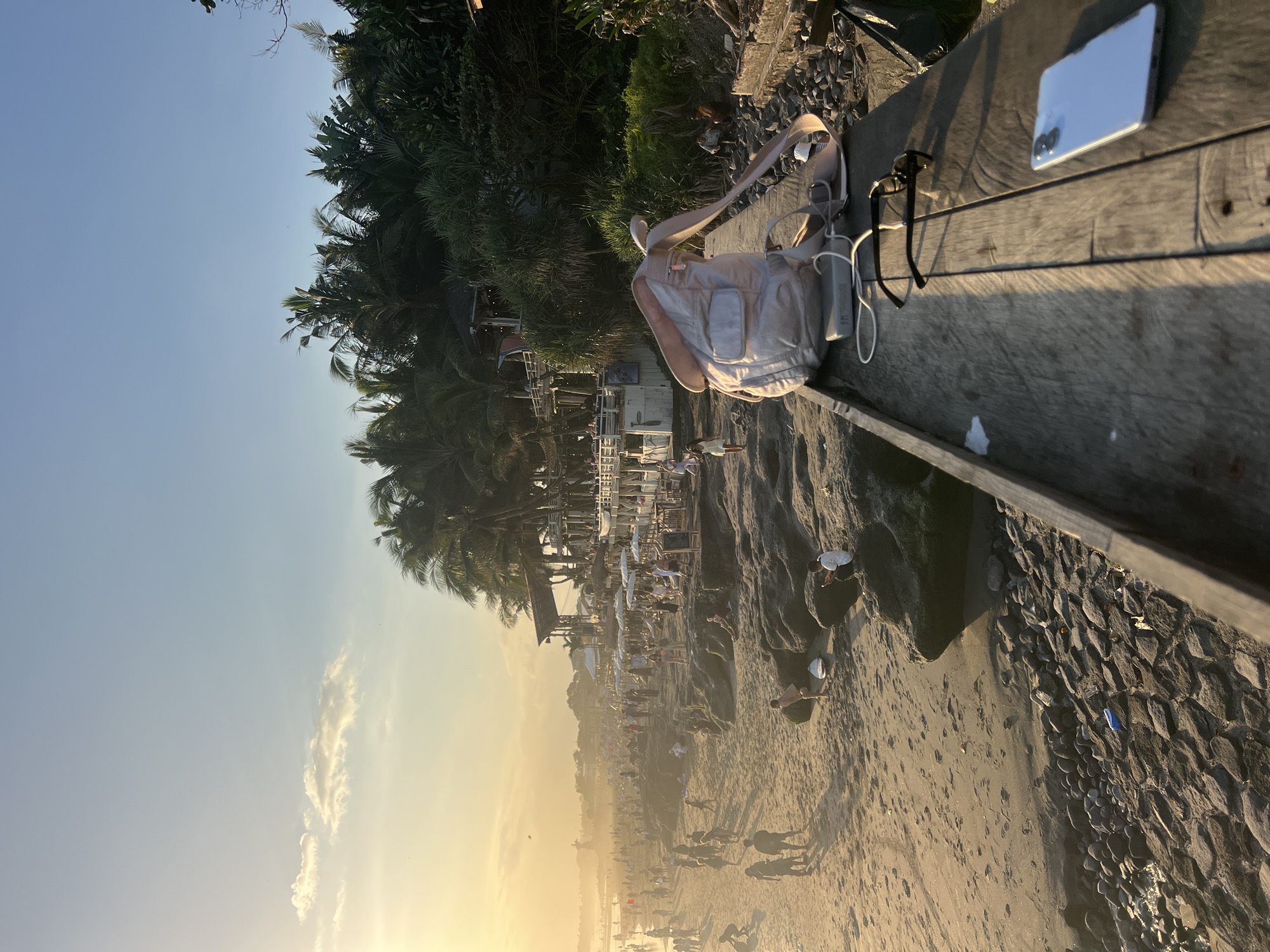 This media file depicts a busy beach scene during sunset in North Kuta, Indonesia. The foreground features a wooden bench with a light-colored bag, a power bank with a white cord, sunglasses, and a white smartphone. The bench overlooks a sandy beach populated with numerous people. In the background, lush green palm trees line the edge of the beach, with several small buildings and thatched-roof structures suggesting a vibrant coastal resort area. The sky is a clear blue with a few wispy clouds, transitioning to a warm, golden hue as the sun sets, casting long shadows across the sand. The overall mood is lively and relaxed, indicative of a popular tourist destination at the end of the day. There is no visible text in the image.