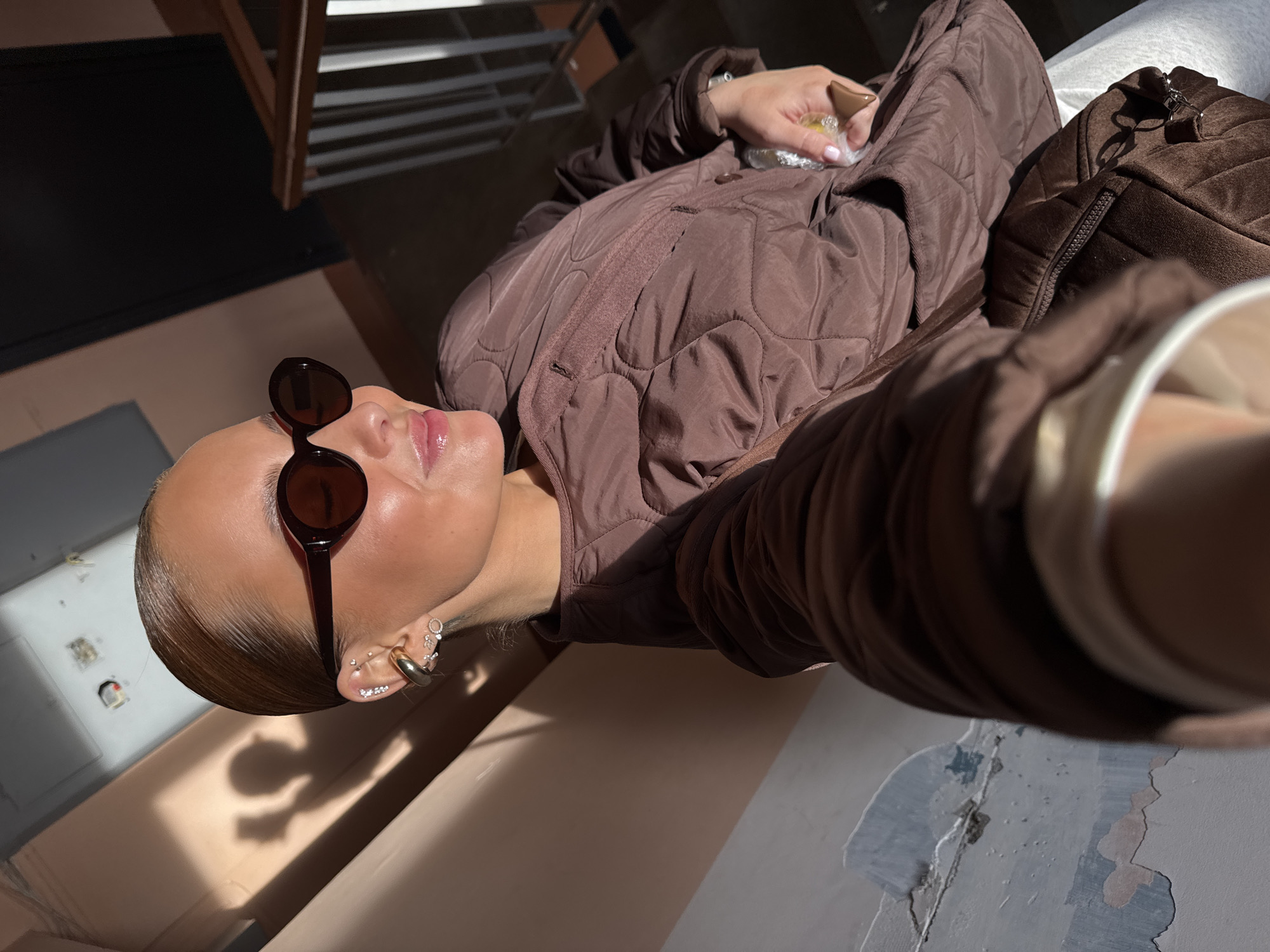 The image depicts a person wearing sunglasses and a brown quilted jacket. They are holding an object wrapped in clear plastic in their hand. The person appears to be in an interior location, possibly an apartment in Chaikovsky, Russia, evidenced by the peeling paint and the presence of interior elements. The perspective suggests the image was taken from a low angle, possibly as a selfie. The image also reveals the person's jewelry, including earrings. A brown corduroy bag is visible next to them.