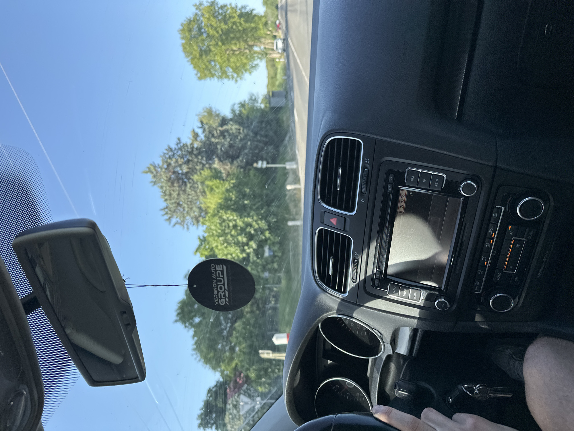 The image is taken from inside a car, looking out through the windshield. A person's hand is visible on the steering wheel, and the car's dashboard with the radio and air conditioning controls is in the foreground. A black car air freshener hangs from the rearview mirror, displaying the text "VERGNON AUTO GROUPE". Outside the car, a tree-lined road is visible under a clear blue sky. The scene appears to be a sunny day in a rural or suburban area. The exact location within Bagard, France, cannot be determined from the visual information alone.