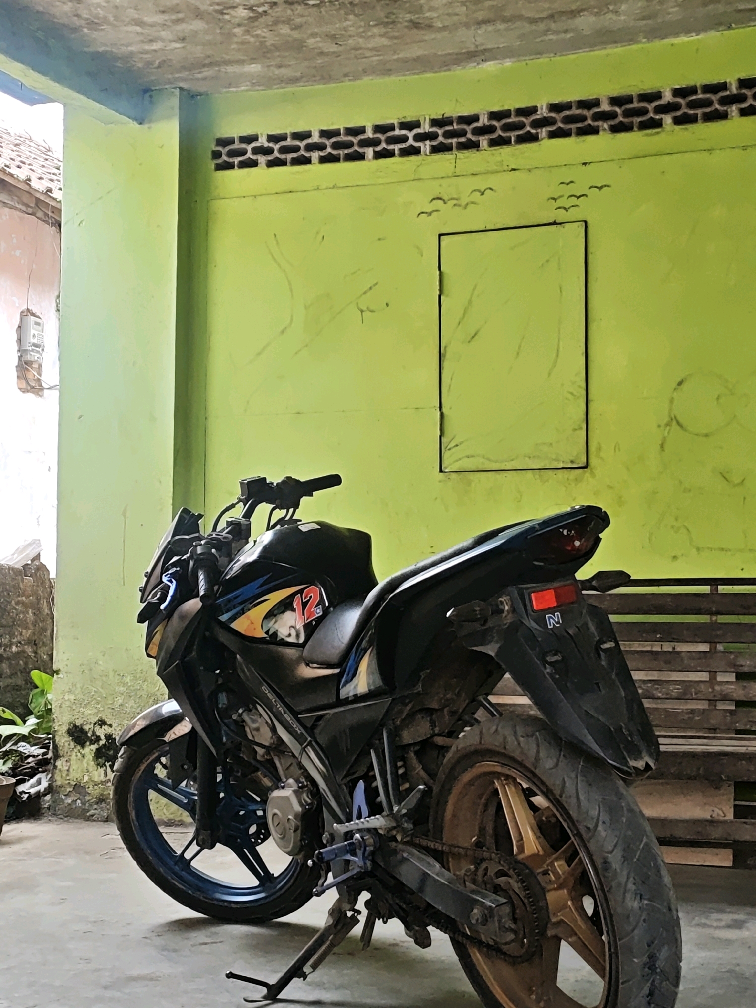 The image depicts a black motorcycle parked on a concrete floor in what appears to be an outdoor sheltered area. The wall behind the motorcycle is painted a vibrant lime green and has some drawn elements, including a rectangular outline and what looks like a flock of birds. Above the green wall is a row of ventilation bricks. To the left of the motorcycle, a portion of a building with a tiled roof and a white electrical meter attached to a pillar is visible. The motorcycle itself is the central focus, with its front wheel turned slightly to the left. It has blue accents on its front wheel rim and a gold-colored rear wheel rim. The rear of the motorcycle features a red taillight and a license plate with the letter "N". To the right of the motorcycle, a wooden structure, possibly a bench or stacked wood, can be seen. The overall scene suggests a residential or semi-commercial outdoor space, possibly within the city of Kemuning, Indonesia, given the architectural elements and the presence of the electrical meter. There are no people visible in the image.