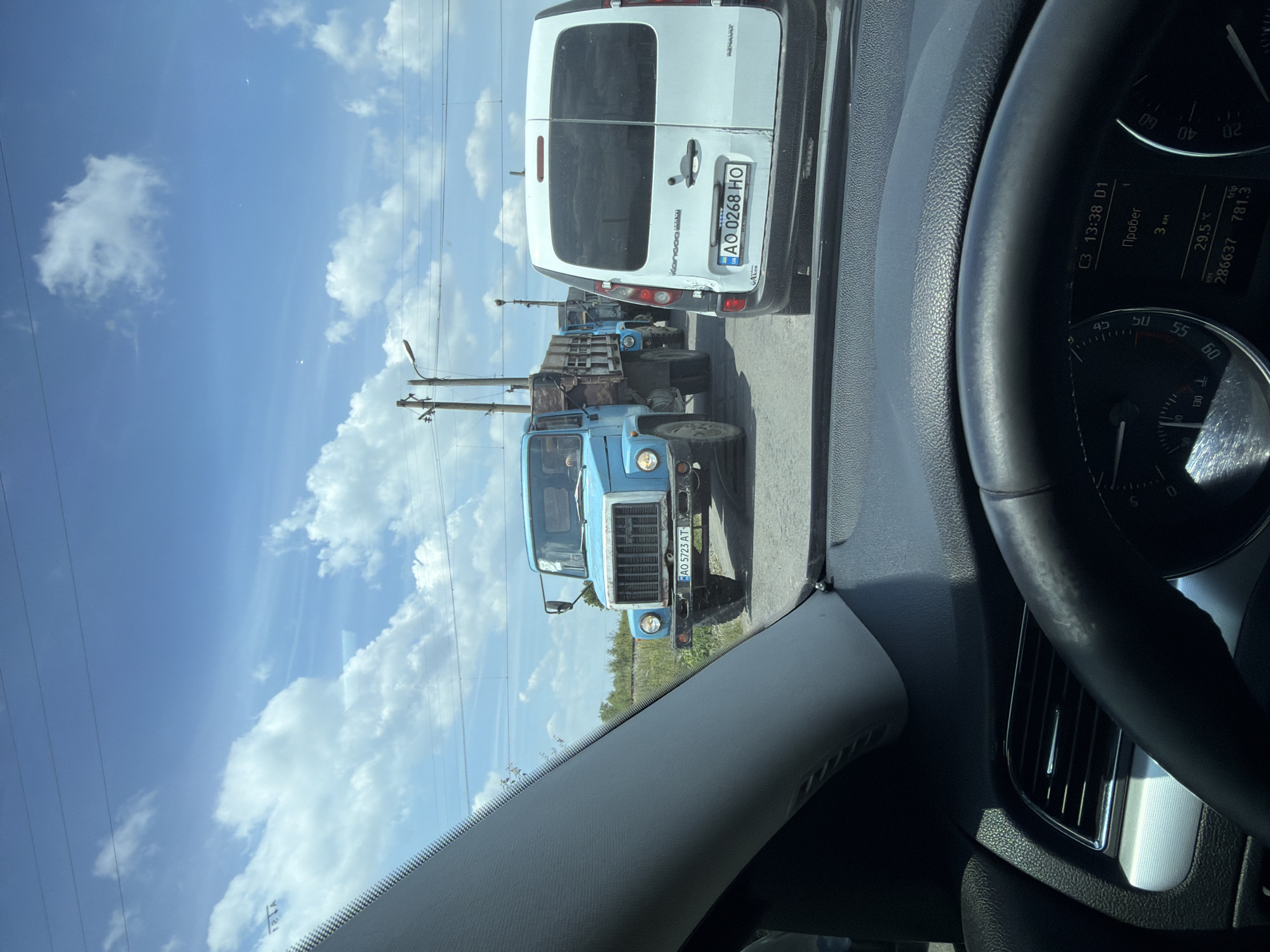 The image shows a view from inside a car, looking forward through the windshield. The car is stopped in traffic on a sunny day. In front of the car, there is a white van, and further ahead, a line of blue trucks. The first blue truck, a flatbed with wooden sides, has the license plate AO 5723 AT. The white van has the license plate AO 0268 HO. The background consists of a blue sky with scattered white clouds, utility poles with wires, and vegetation. The location is identified as Tarnivtsi, Ukraine, based on the provided context. The dashboard of the car is partially visible in the foreground, showing a speedometer and a digital display with time, temperature, and mileage.