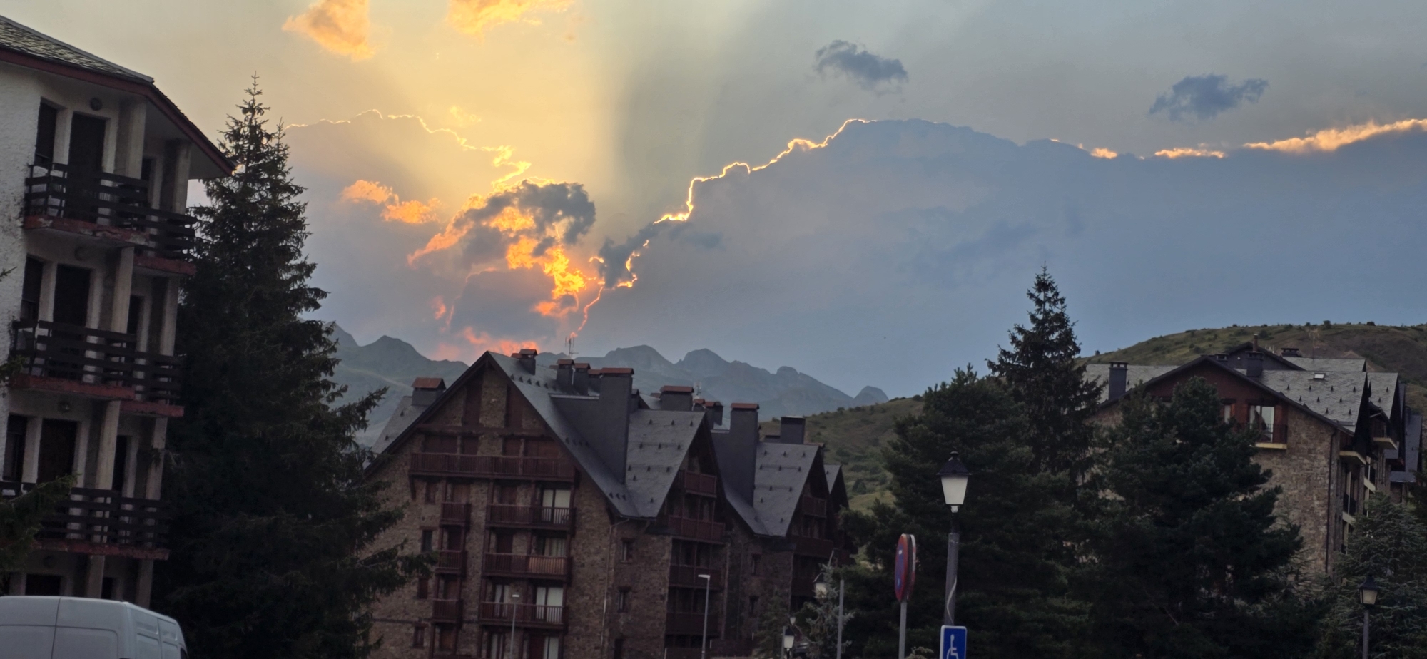 The image depicts a mountainous landscape in Sallent de Gállego, Spain, under a dramatic sunset. The sky is filled with vibrant orange and yellow clouds, with rays of light piercing through them. In the foreground, several multi-story buildings with dark roofs and balconies are visible, interspersed with tall evergreen trees. The architecture suggests a ski resort or mountain village setting. In the distance, rugged mountains form a silhouette against the colorful sky. The overall scene is one of natural beauty enhanced by the atmospheric conditions of a sunset.
