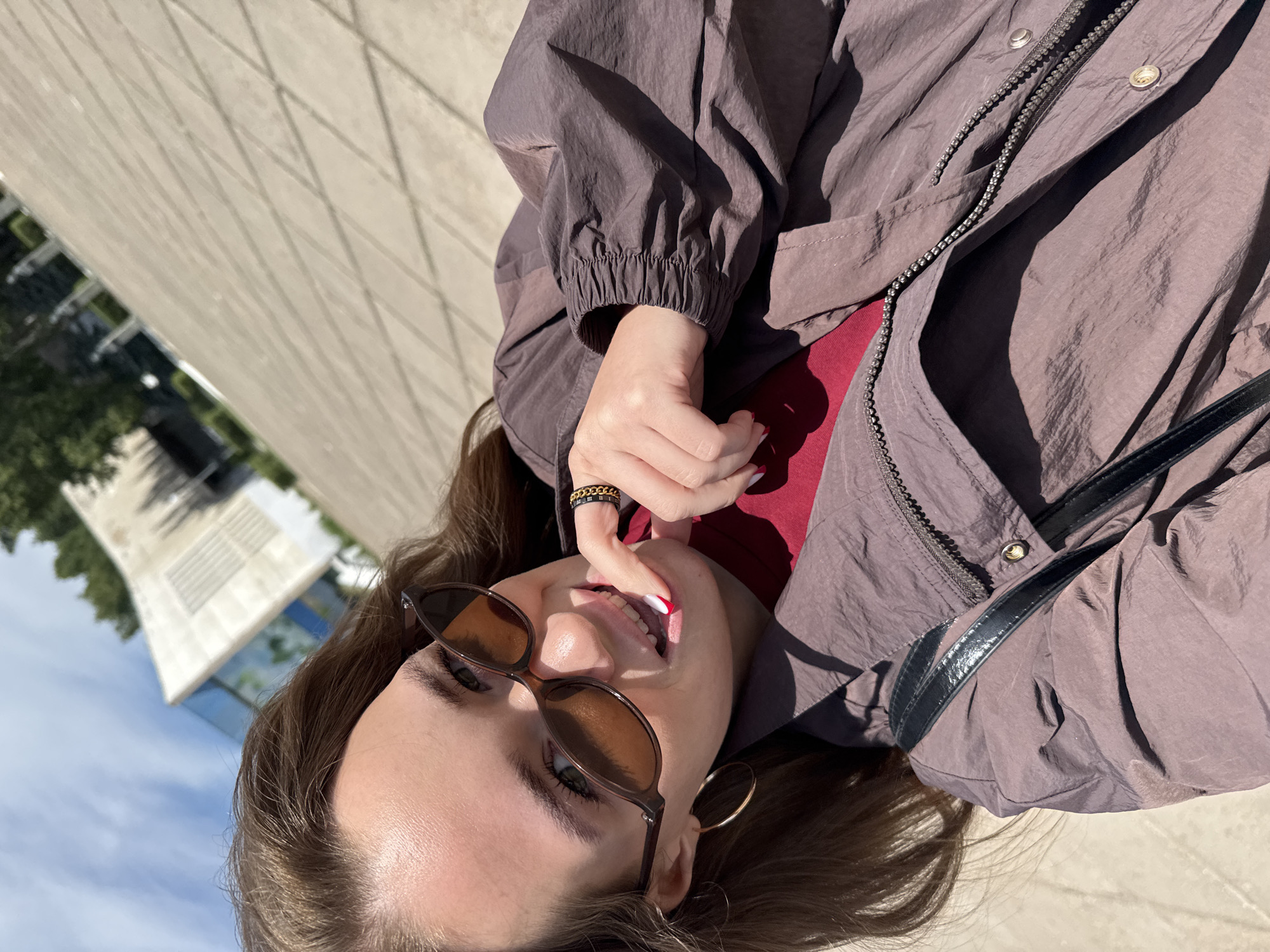 In a daylight, selfie-style shot, a young woman with brown hair, wearing sunglasses and a reddish shirt under a purple jacket, poses outdoors on a paved area. She has her right index finger near her mouth, showing red and white nail polish. A black purse strap is visible across her shoulder. In the background, a modern building with large glass windows and trees can be seen. The setting is likely an urban environment, possibly Chaikovsky, Russia, given the architectural style and general appearance of the locale.