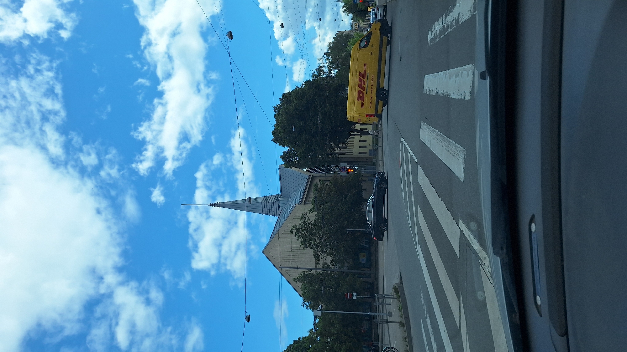 The image captures a street scene in Riga, Latvia, featuring a building with a distinctive spire, possibly a church or public building, dominating the background. In the foreground, a DHL delivery van is visible in motion, alongside a black sedan on the road. The intersection is marked with pedestrian crossings, and traffic lights are present. The sky is blue with scattered white clouds. The perspective suggests the photo was taken from within a vehicle.