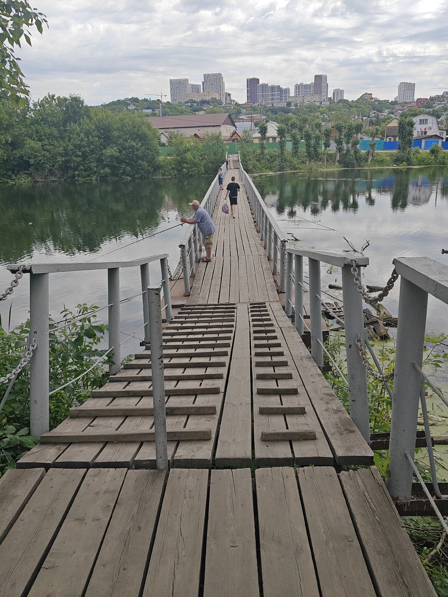 An eye-level, medium shot shows a wooden pedestrian bridge extending over a calm body of water. The bridge, constructed of weathered planks, has metal railings with chains for support. On the bridge, a man in a light-colored hat and blue shirt is fishing, holding a rod over the water. Further down the bridge, a person in a black shirt and shorts walks away from the camera, carrying a bag. In the background, a city skyline with modern buildings rises behind a line of trees and houses. The sky is overcast. The water reflects the surrounding trees and buildings. The overall scene depicts a serene moment of leisure in an urban-natural setting.