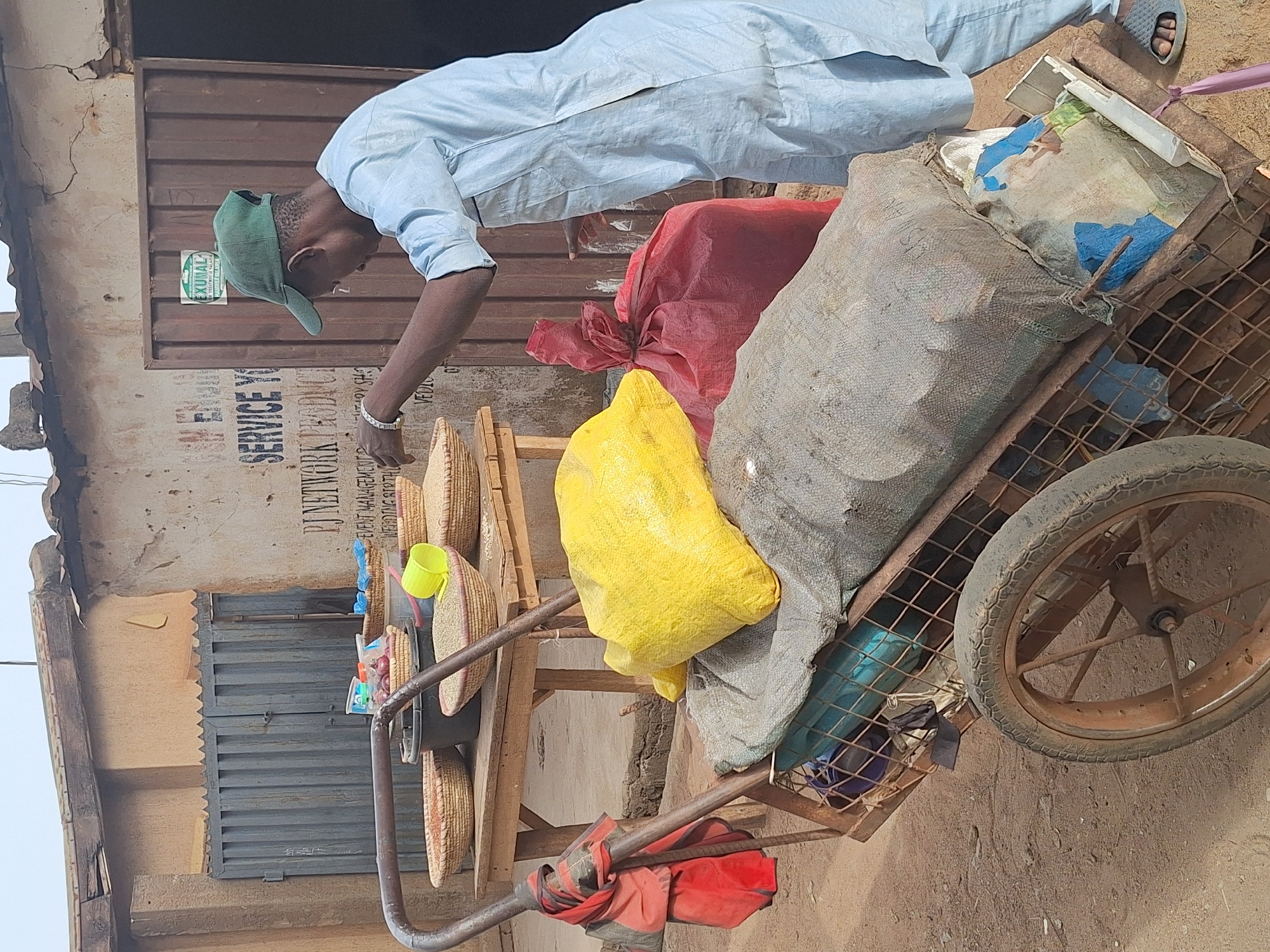 A man is standing over a cart full of items, possibly merchandise for sale. The man is wearing a light blue long-sleeved shirt, a green baseball cap, and sandals. He has a bracelet on his left wrist. The cart is loaded with items in red, yellow, and gray bags, as well as woven baskets. The cart's wheel is visibly worn and dirty. The scene is set outside, with a building in the background that has a partially obscured sign with the text "PJ NETWORK" visible. Based on the provided location, this scene is likely taking place in Sabon Tasha, Nigeria.