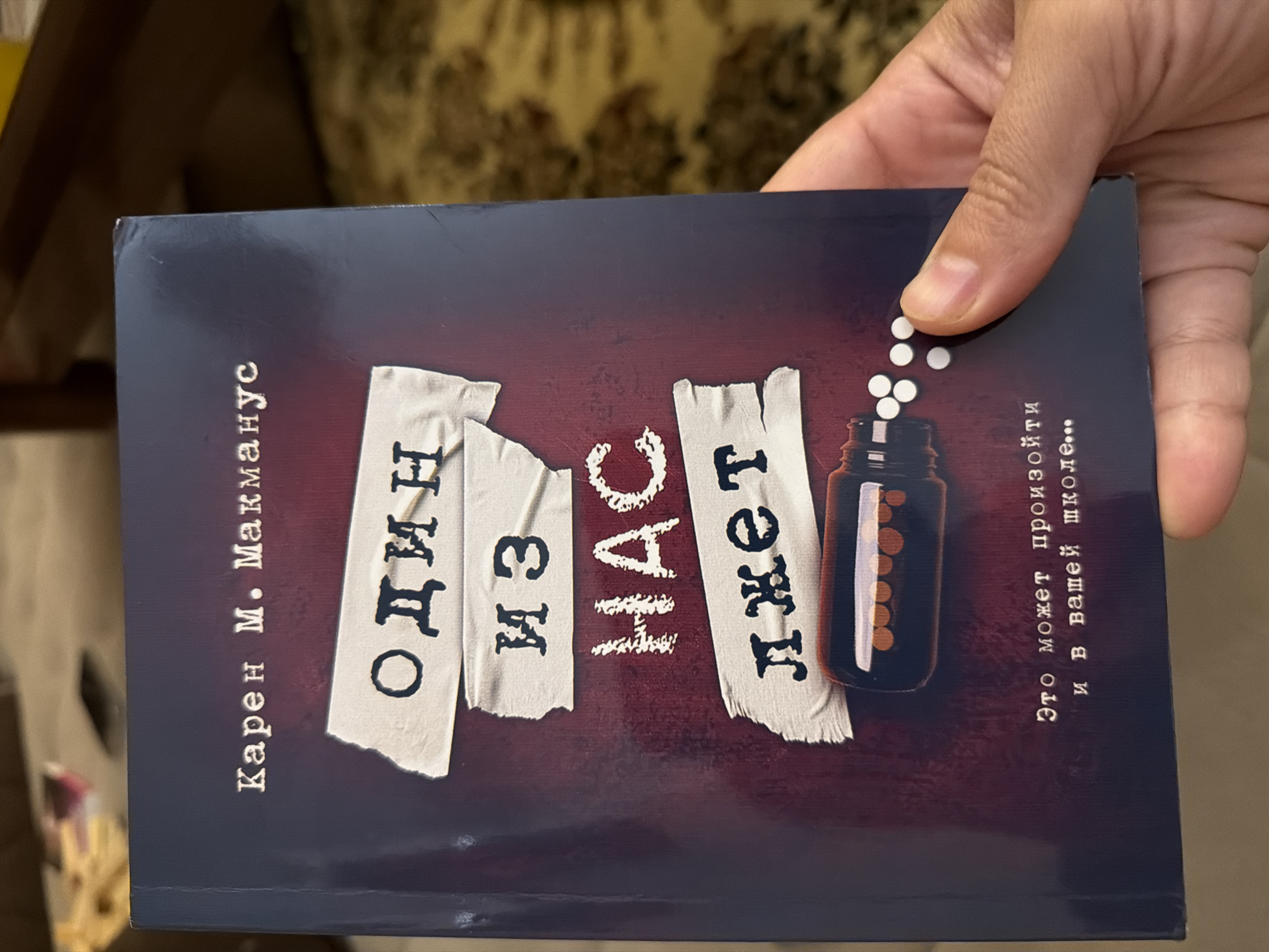 The image shows a close-up of a book cover held by a person's hand. The book is dark blue with a distressed, deep red background. The title, "ОДИН ИЗ НАС ЛЖЕТ" (One of us is lying), is printed in large, distressed white letters on pieces of tape that are adhered to the cover. Above the title is the author's name, "Карен М. Макманус" (Karen M. McManus), in smaller white serif font. At the bottom of the cover, there is an illustration of a pill bottle with pills and scattered white pills. Below the illustration, a tagline reads, "Это может произойти и в вашей школе..." (This can happen in your school too...). The background of the image is blurred, showing what appears to be a patterned carpet and furniture. The image is a still photograph and depicts no recognizable landmarks or features that would place it in Koshkurgon, Uzbekistan.