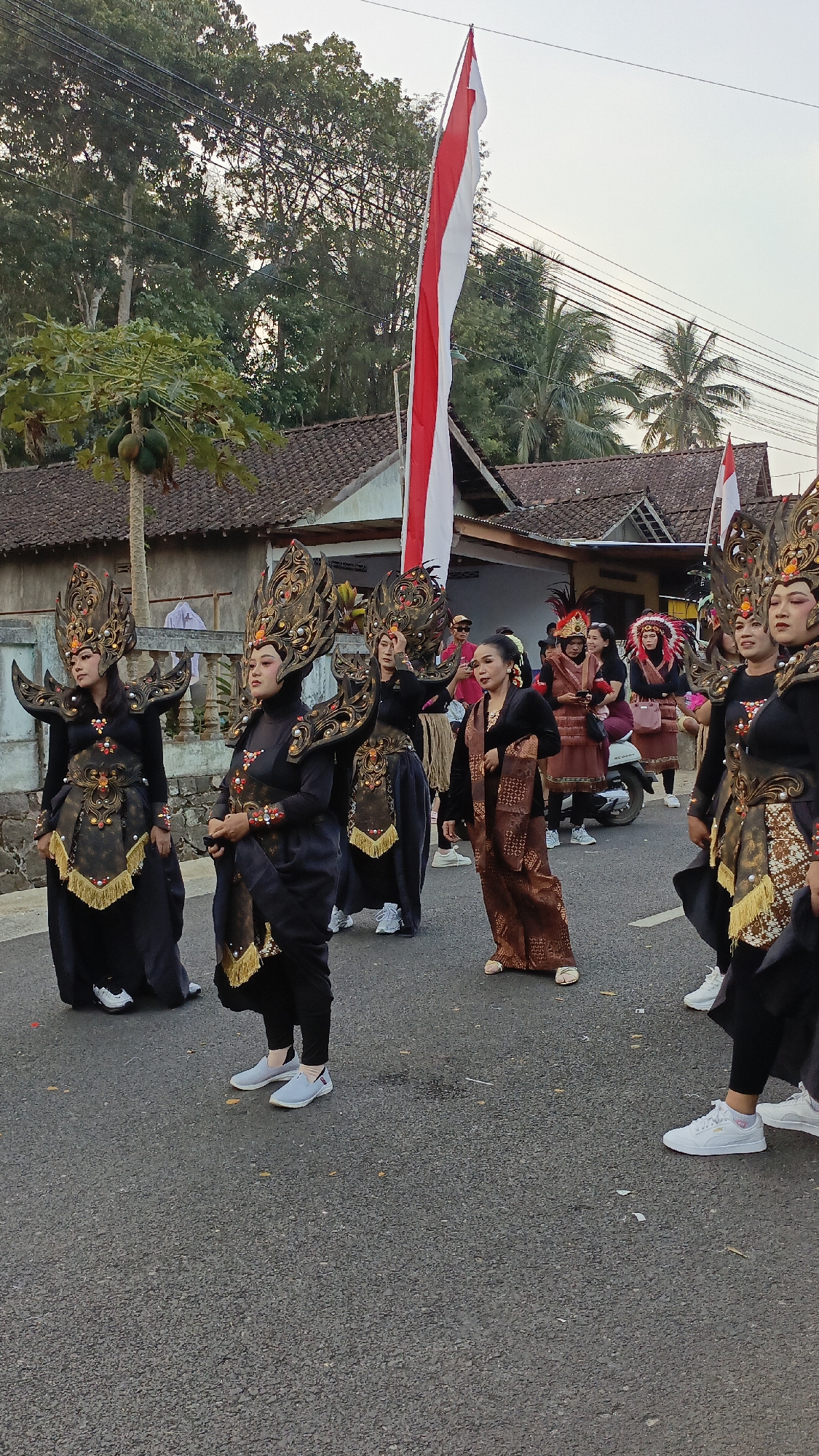The image captures a street scene in Krajan Lor, Indonesia, featuring a procession of people in traditional attire. In the foreground, several women are walking, dressed in elaborate black costumes adorned with gold embellishments and headdresses. One woman in the center wears a brown patterned sarong and a black top. Behind them, other participants are visible, some wearing headdresses reminiscent of Native American war bonnets, and one person is riding a scooter. A tall Indonesian flag, red and white, is prominently displayed, waving in the background. The environment includes buildings with tiled roofs, trees, and utility poles with visible wires, indicating a rural or semi-urban setting. The overall atmosphere suggests a cultural event or celebration.