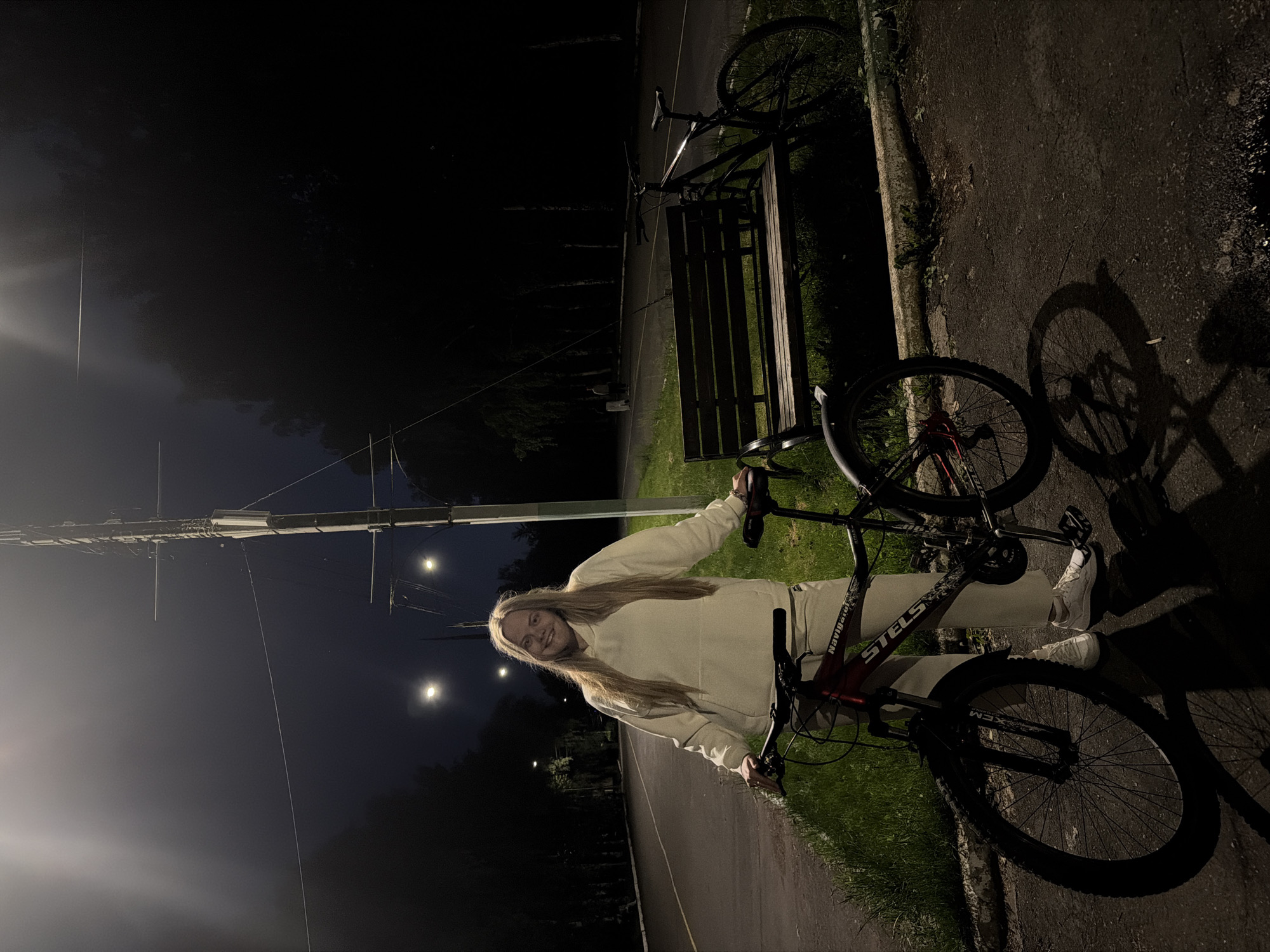 On an asphalt path at night in Izhevsk, Russia, a young woman with long blonde hair stands smiling widely, holding the handlebars of a bicycle. She wears a light-colored, long-sleeved hooded top, matching pants, and white sneakers. The bicycle she is with is dark-colored with red accents; the brand "STELS" is visible on its frame, along with "Navigator" and "XCM" on the front fork.
The setting is an outdoor area, likely a park, illuminated by streetlights that cast long shadows, including a distinct bicycle shadow on the path. To her left, the asphalt path extends, bordered by grassy areas and dark trees. Further back and to the left, another bicycle is parked next to a wooden bench. The sky is dark, indicating nighttime, with a few bright streetlights visible in the distance. The woman's joyful expression contributes to a pleasant and relaxed atmosphere.