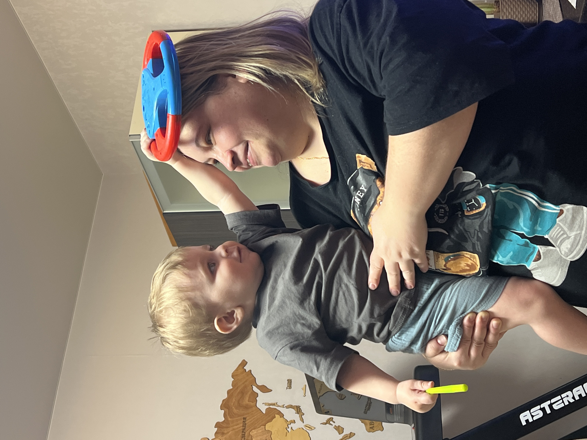 A woman in a black T-shirt holds a young child in her arms. The child is wearing a gray T-shirt and blue shorts. The woman is smiling at the child, who is looking up at a blue and red toy steering wheel that the woman is holding above him. The child is also holding a yellow crayon.
A wooden map of the world is visible on the wall behind them. The letters "ФЕДЕРАЦИЯ" and "РОССИЯ" are visible on the map, which indicates that the location may be in Russia. The city of Perm is not directly discernible from the image.