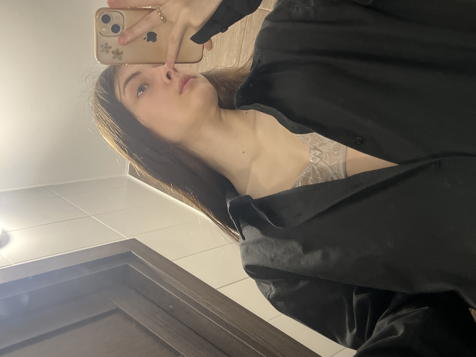A young woman is taking a selfie in what appears to be a bathroom with white tiled walls. She is holding a gold-colored iPhone with a floral design on the case. She is wearing a black, buttoned-down shirt that is slightly open, revealing a lace bra. The background is a white tiled wall with a wooden-framed door. The location is not identifiable as Saint Petersburg, Russia.