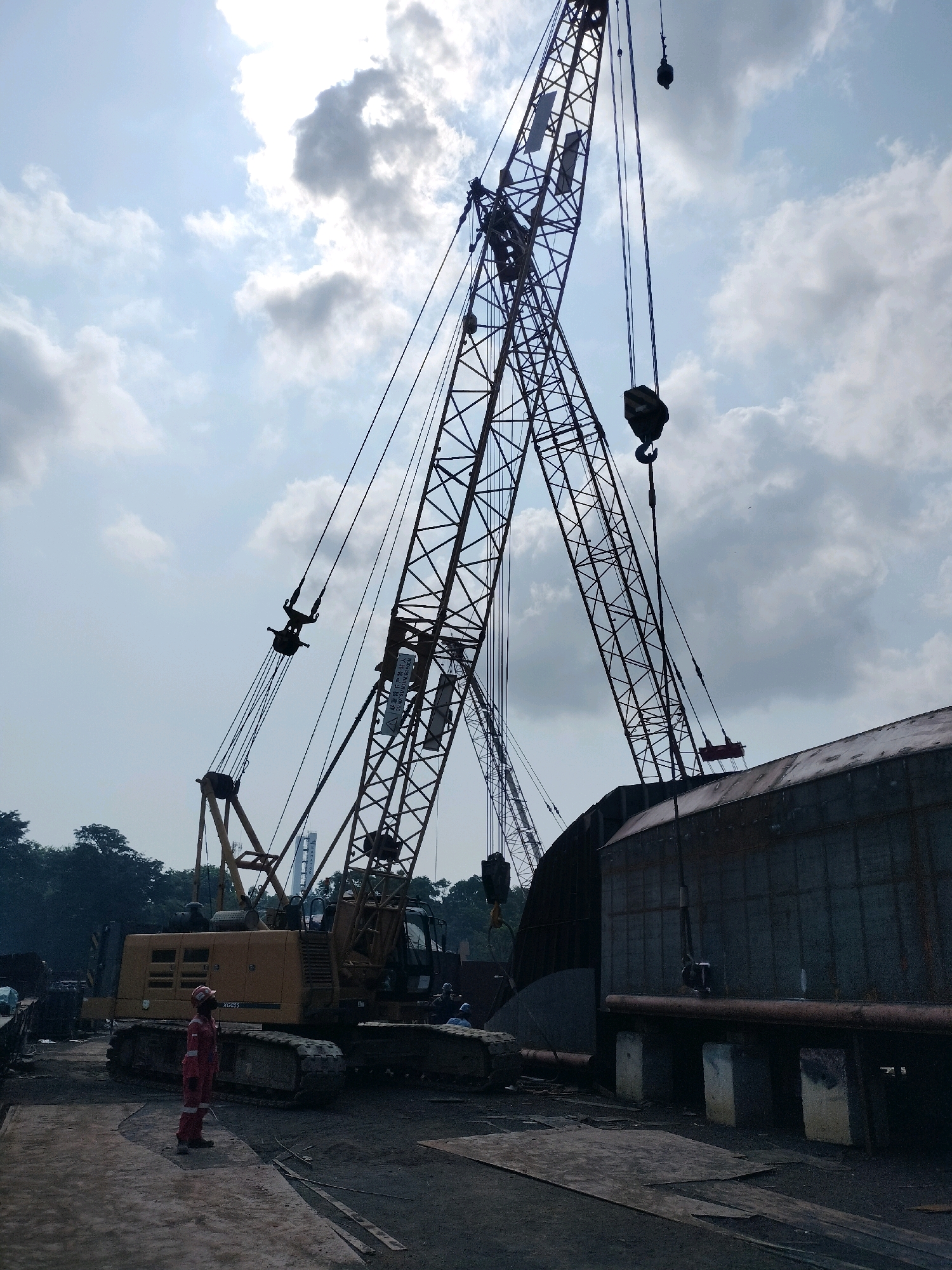 The image captures a construction or industrial scene, likely in Batam, Indonesia, given the context of shipbuilding or port operations. Dominating the frame are two large cranes, one yellow and tracked, the other a taller lattice boom crane, both silhouetted against a cloudy sky. The tracked crane appears to be a Caterpillar model, identified by the "XGC55" marking on its side. A person in a red hard hat and jumpsuit stands on the ground, facing towards the cranes, seemingly supervising or observing the work. In the foreground, a large, curved metal structure, possibly part of a ship or industrial building, is under construction. The ground is rough and unpaved, with scattered metal plates and debris, indicating an active work area. The overall atmosphere is one of heavy industry and ongoing construction.