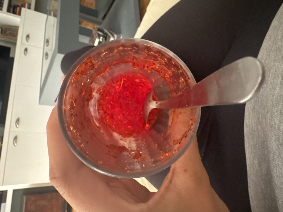 A first-person overhead view depicts a clear glass held by a hand over a person's lap in an indoor setting in Mexico City, Mexico. The glass contains remnants of a reddish-orange granular substance adhering to its inner walls and a dense, bright red, gel-like or spherical material at its base. A silver metallic spoon is inserted into the glass, its bowl submerged in the red material. The hand holding the glass is positioned on the left side. Below the glass, dark fabric, likely leggings or pants, covers the individual's thighs, with a lighter grey fabric visible beneath. The blurred background reveals a white cabinet or shelving unit with silver handles, and to its right, a dark grey rectangular table with a reflective glass top, suggesting a domestic environment.