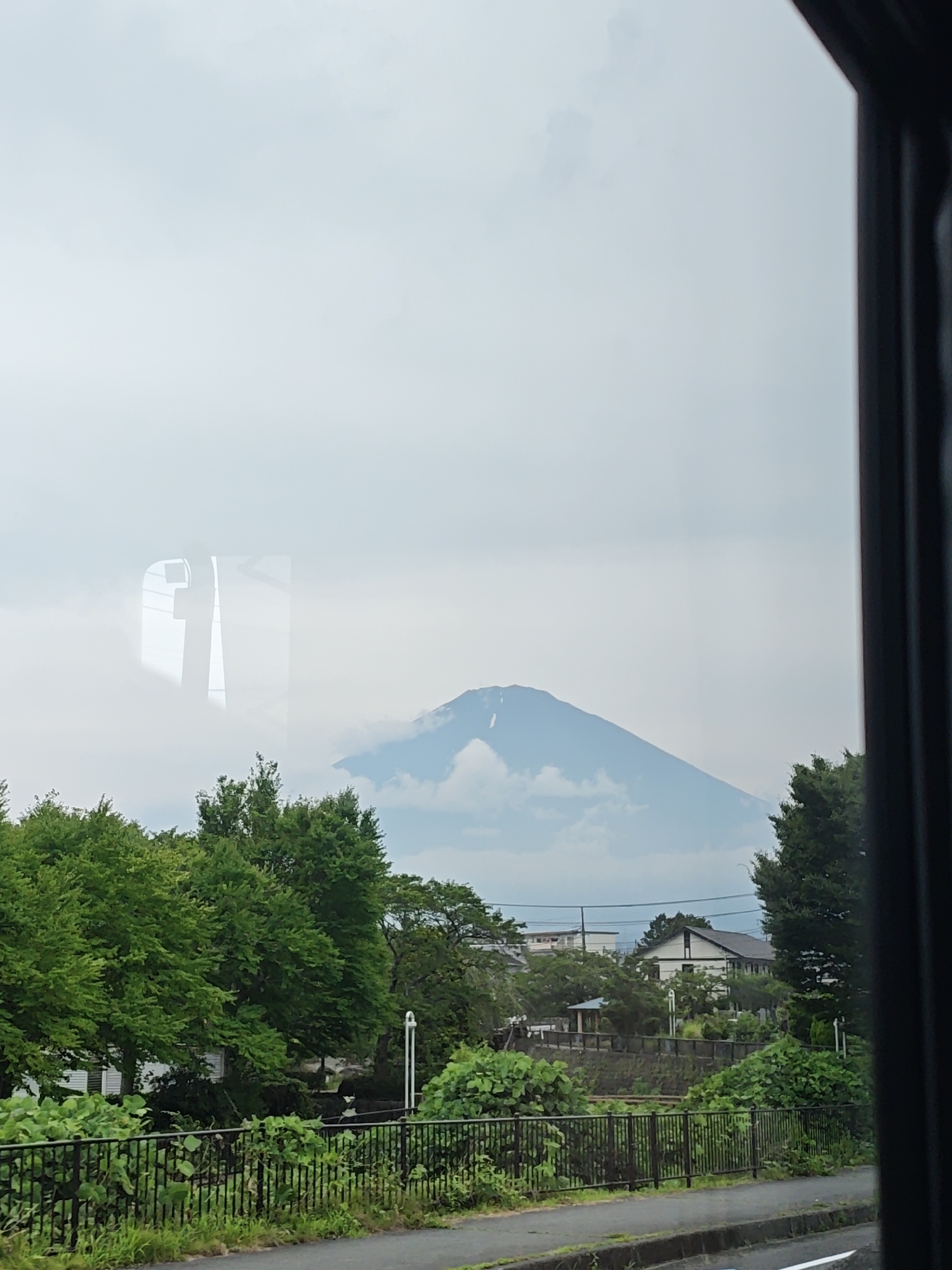 The image depicts a view of Mount Fuji, partially obscured by clouds, from the city of Gotemba, Japan. The foreground features lush green trees lining a black metal fence, with a paved road visible beyond. In the mid-ground, residential buildings and more trees are scattered. The sky is overcast, contributing to the soft lighting of the scene. The reflection of what appears to be a window frame is visible on the mountain itself.