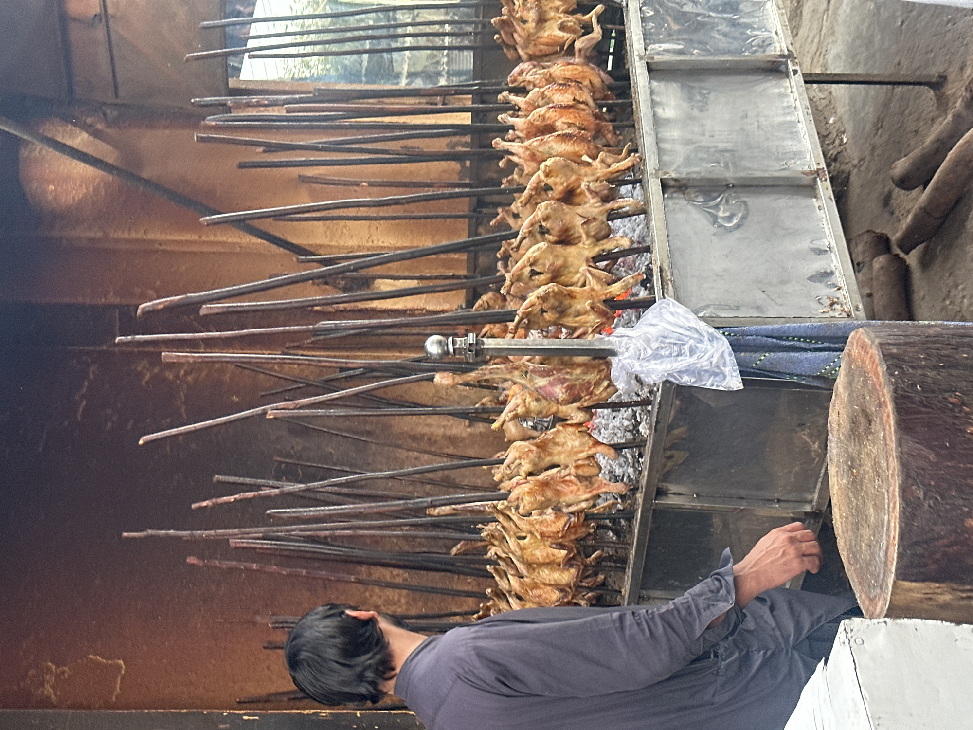 A street vendor in Quetta City Tehsil, Pakistan, tends to numerous chickens skewered on metal rods over hot coals. The chickens, arranged in rows, cook over the fire. The vendor, dressed in a dark long-sleeve shirt, is focused on the cooking process. A metal drip pan sits beneath the chickens, collecting rendered fat and juices. A large log is positioned to the right of the cooker.