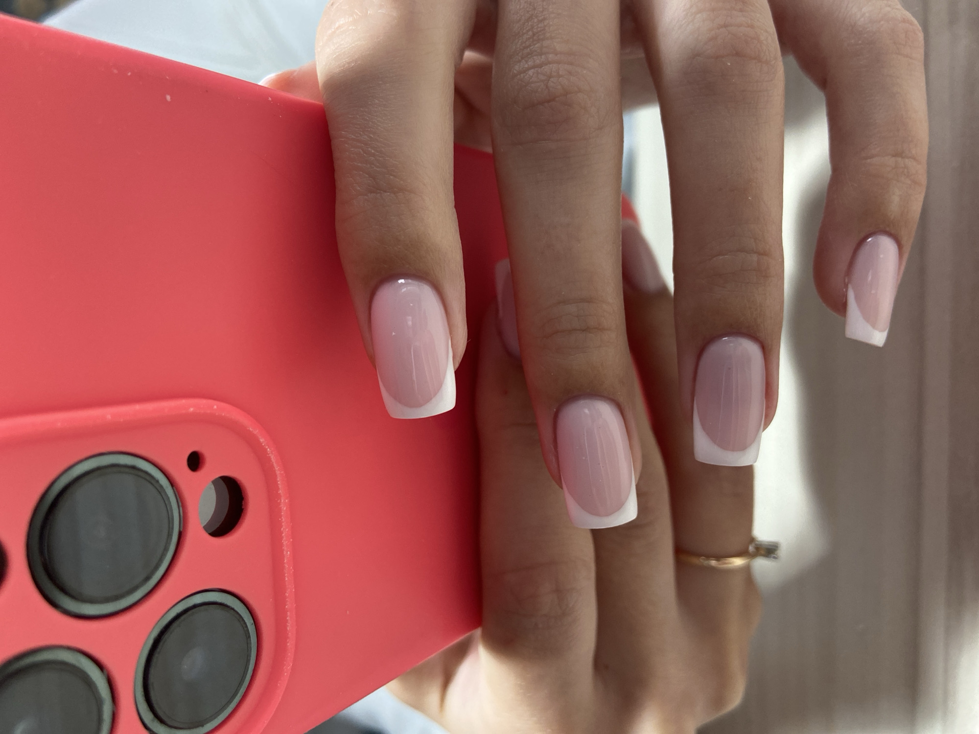A person's hand is holding a red smartphone with visible camera lenses. The hand has manicured nails with a French tip design. A gold ring is visible on one finger. The background is a blurred white surface. The location is not discernable from the image.