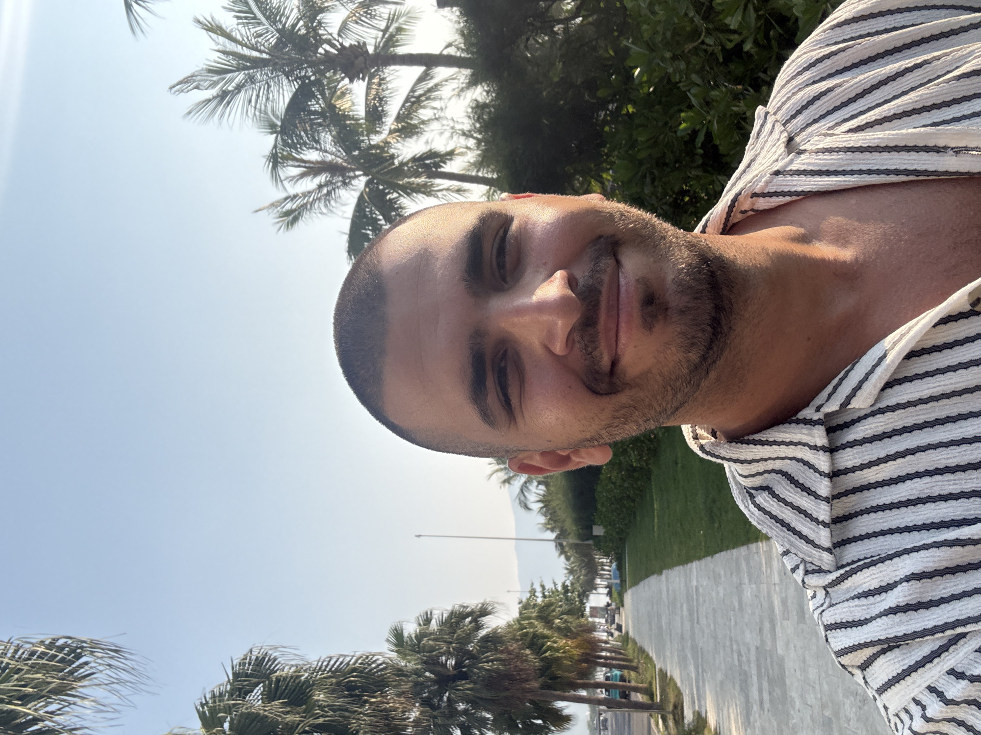 A man with a shaved head and a trimmed beard smiles at the camera. He wears a white striped shirt. Behind him is a street lined with palm trees. Beyond the trees is a glimpse of the ocean. This is likely in Đà Nẵng, Vietnam, a coastal city known for its beaches and palm-lined streets.