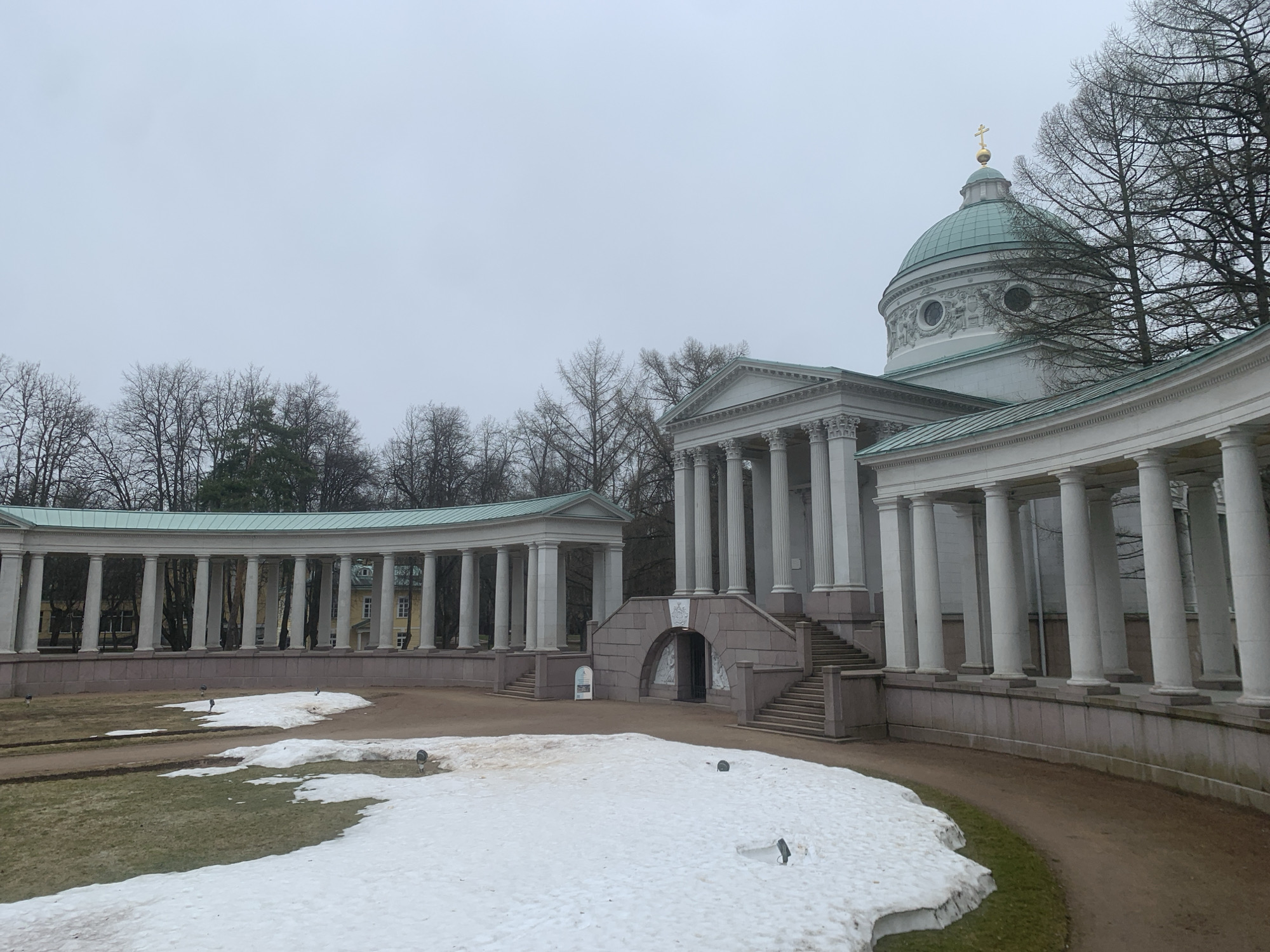 The image shows the Colonnade and Mausoleum of the Yusupov family in Arkhangel’skoye, Russia. The colonnade is a curved structure with white columns, and the mausoleum is a classical building with a dome topped by a golden cross. The ground is partially covered with snow. The sky is overcast. Bare trees are visible in the background.
