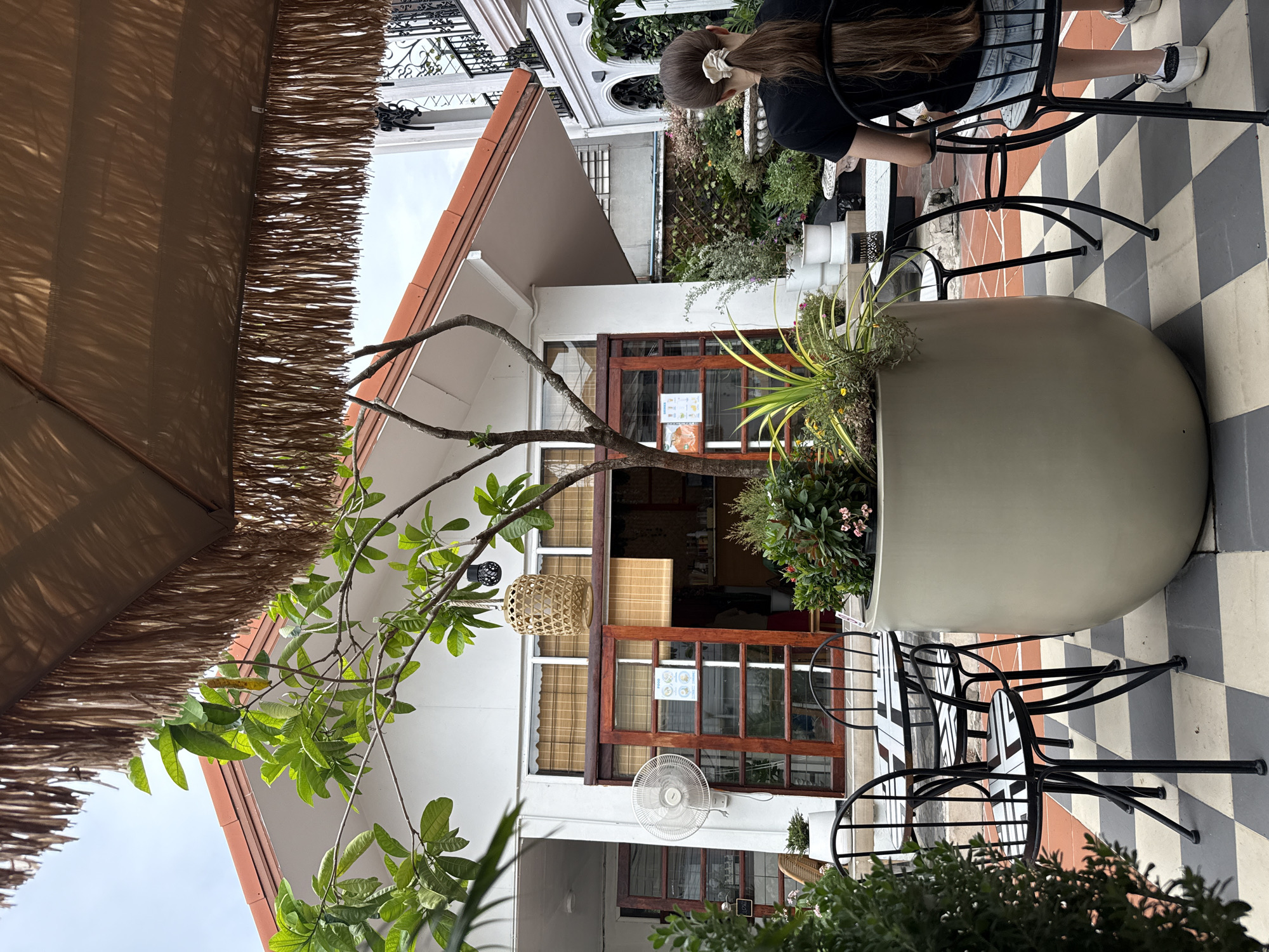 An outdoor seating area of a cafe is depicted in Bangkok, Thailand, under a large thatched umbrella. A person with long brown hair tied in a ponytail, wearing a black t-shirt and denim shorts, is seated at a table. The table and chairs have a black and white checkered pattern. In the foreground, a large beige planter overflows with greenery and small pink flowers. Behind the planter, a tree with bare branches and green leaves reaches towards the sky. The cafe building, painted white with orange trim, features large wooden-framed windows and doors. A white electric fan is mounted on the wall. The ground is covered in a black and white checkered tile pattern. To the right, a white building with ornate black railings is visible, suggesting an alleyway or a connected structure. The overall ambiance is relaxed and inviting, with lush vegetation and charming architectural details.