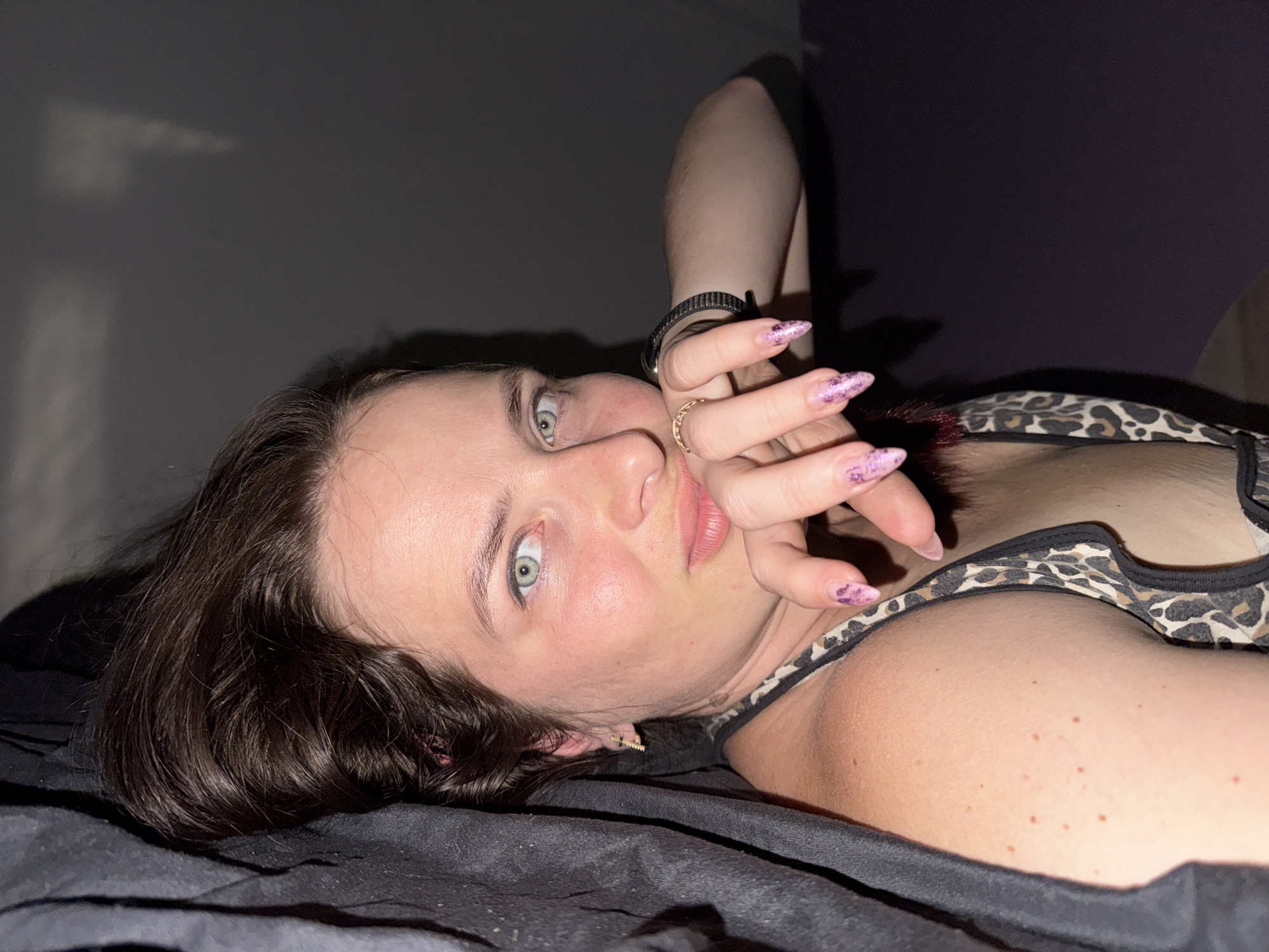 A young woman lies on her back on a black surface, likely a bed, with her head turned towards the viewer. She has long, dark, wavy hair that frames her face and spills onto the surface. Her eyes are wide open and a striking light blue-green, looking directly at the camera. Her left hand is raised, with manicured nails adorned with pink glitter, and her fingers are interlaced across her mouth and nose area. She is wearing a leopard print top. The background is dark and out of focus, with a faint shadow or light pattern on the wall behind her. The lighting suggests an indoor setting, possibly a bedroom, with some artificial light creating shadows. No specific city or location details are evident.