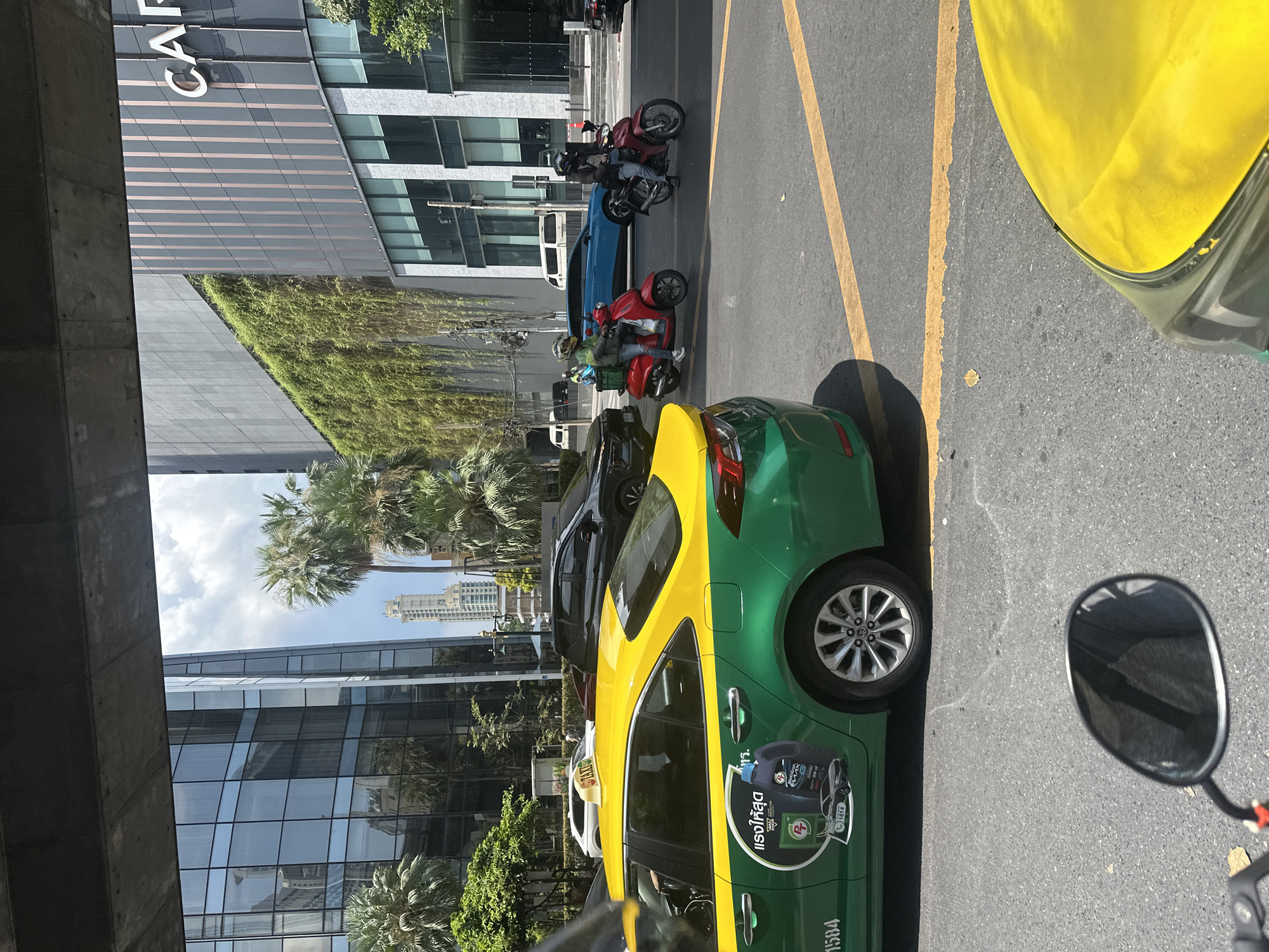 The image captures a street scene in Bangkok, Thailand. A green and yellow taxi cab is prominently featured, partially obscuring the view to the right. Several parked motorcycles and a blue car are visible in the lane adjacent to the taxi. The background includes a modern building with a glass facade and some greenery. Palm trees and a partial view of the Bangkok skyline, including a tall building, can be seen in the distance. The photograph is taken from an elevated position, as evidenced by the concrete structure in the lower left. The scene suggests a busy urban environment with various modes of transportation and commercial buildings.
