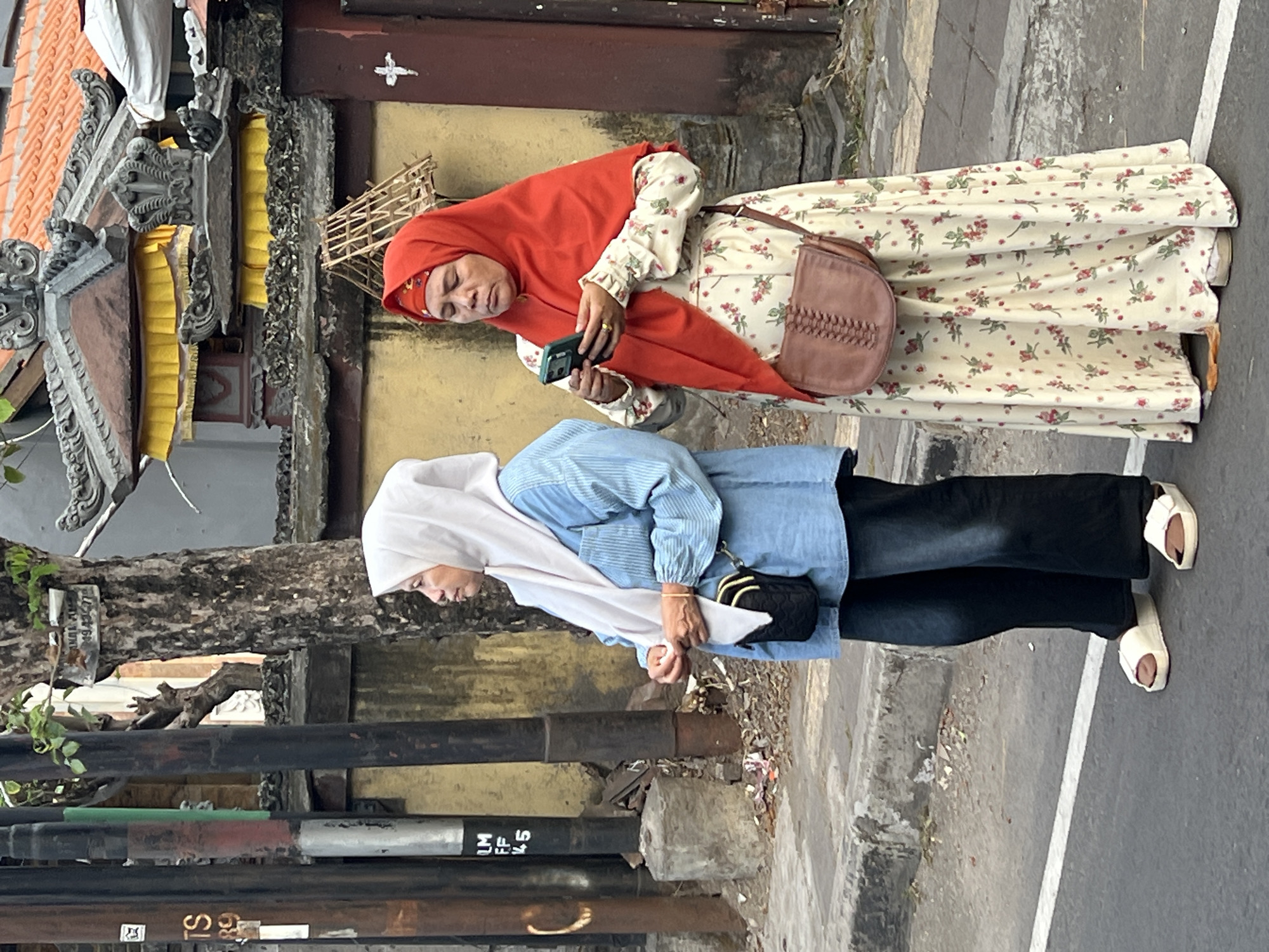 Two women, dressed in hijabs and modest clothing, stand on a paved street with a sidewalk and a textured yellow wall behind them. The woman on the left wears a white hijab and a light blue button-down shirt over dark pants and white slip-on sandals. She has a small black crossbody bag. The woman on the right wears a bright red hijab and a cream-colored floral print dress with a brown woven crossbody bag. She is holding a smartphone in her hands and looking at it. The background includes a traditional Balinese architectural structure with layered roofs and yellow fabric drapes, along with several utility poles. Debris is visible on the sidewalk. The scene appears to be in Denpasar, Indonesia, suggested by the architectural style and the presence of utility poles common in urban Indonesian settings.