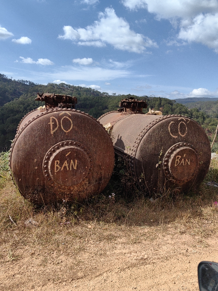 The image displays two heavily rusted, derelict cylindrical metal tanks or boilers resting on dry, sparse grass and reddish-brown dirt. The foreground includes patches of dry grass, small green weeds, and bare earth. The tank on the left, more prominent, features the text "ĐỒ BÁN" (Item for Sale) painted in faded yellow on its circular end face. The partially visible tank on the right displays "CƠ BÁN" (Mechanism for Sale) in similar paint on its end face. Both tanks exhibit significant surface corrosion, visible rivets along their sides and circular end panels, and decayed structures on their top surfaces. In the background, a deep valley separates the immediate location from distant, densely forested green hills under a clear blue sky with scattered white cumulus clouds. A faint amusement ride, possibly a Ferris wheel, is discernible on a distant hilltop to the right. No individuals are present. The scene is located in Păng Tiêng 1, Vietnam.