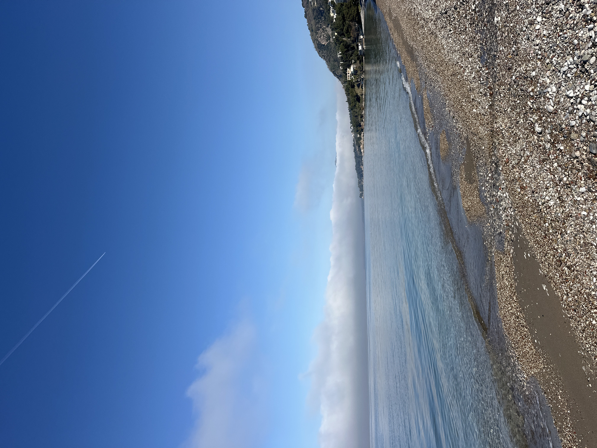The image shows a pebble beach in Šušanj, Montenegro. The Adriatic Sea is on the right, meeting the shoreline. A line of vegetation is above the beach, with buildings visible in the distance on a hill. The sky is clear blue with some clouds in the lower part of the frame. A contrail of an airplane is visible in the sky on the left side.