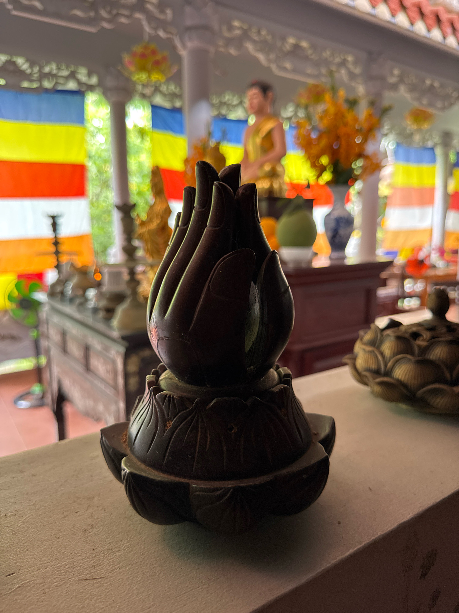 The image depicts a close-up view of a dark, intricately carved incense burner shaped like a lotus flower, resting on a light-colored ledge. In the background, out of focus, is an indoor setting that appears to be a Buddhist temple in Nha Trang, Vietnam. Various prayer flags in vibrant colors (red, yellow, blue, orange) are visible, strung across the space. A blurred figure of a person in a golden robe, likely a Buddha statue, is present behind the incense burner. Several other decorative objects, including vases and what appear to be other incense burners, are also visible in the background. The overall lighting suggests it is daytime.