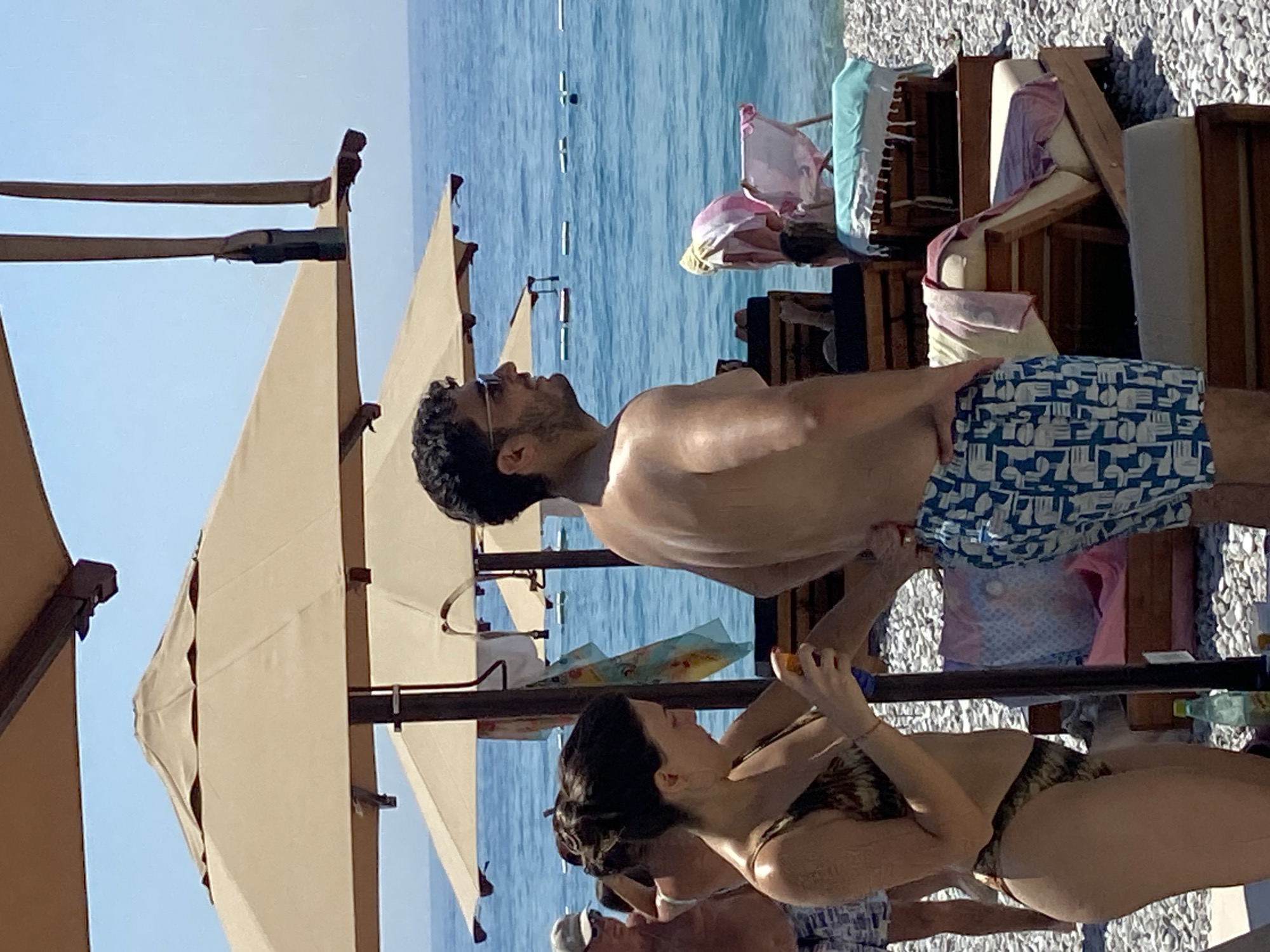 A medium shot shows a woman in a bikini applying sunscreen to her arm with a spray bottle on a pebble beach near the city of Prijevor, Montenegro. She stands next to a man in patterned swim trunks. In the background, the turquoise sea meets a clear blue sky. Several beach umbrellas and lounge chairs are visible, some draped with towels.
