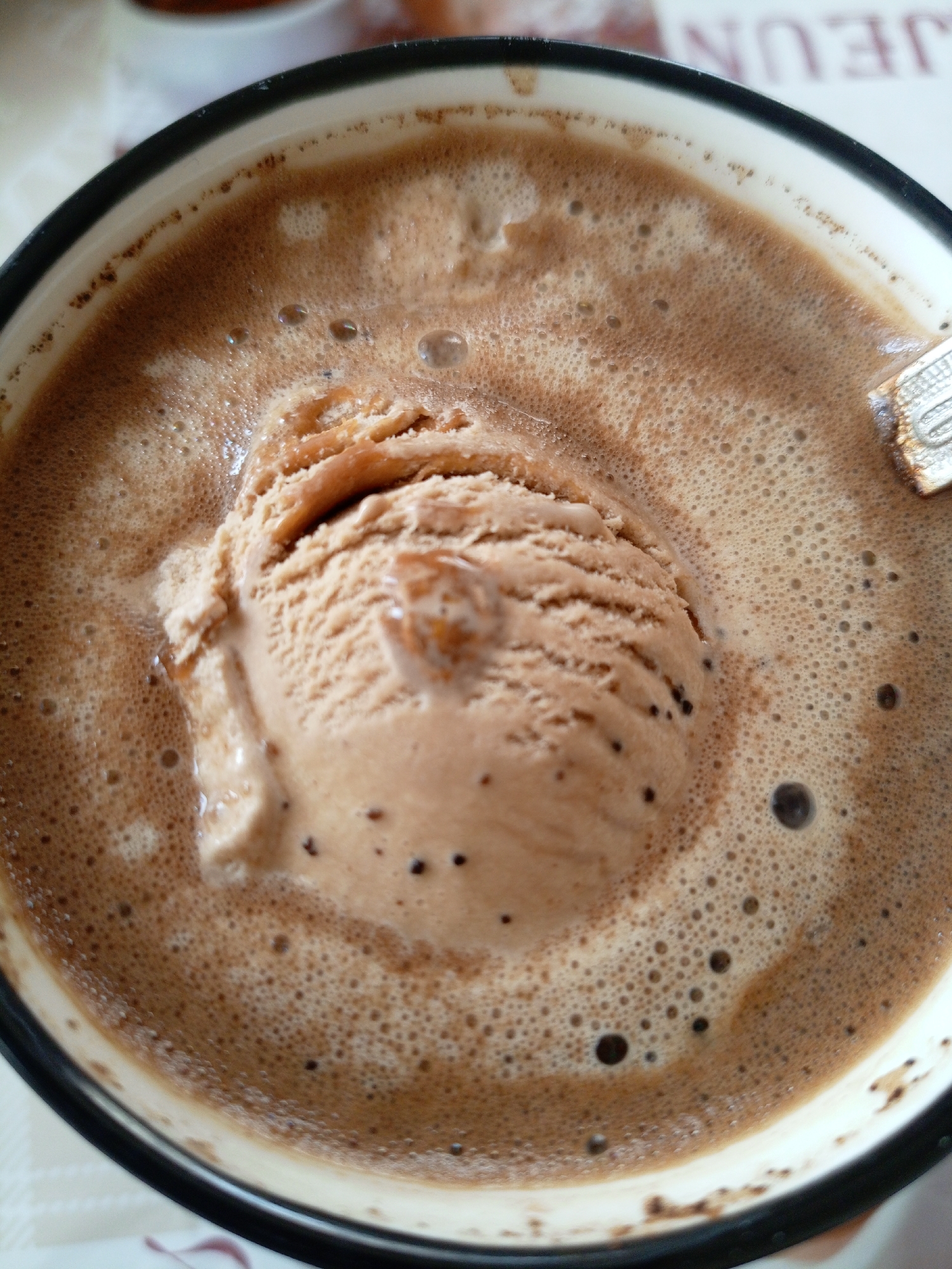 An overhead, close-up photograph depicts a single scoop of light brown, possibly chocolate or coffee-flavored, ice cream partially submerged and melting into a dark brown, frothy liquid contained within a white ceramic or enamel cup with a prominent dark rim. The liquid surface is covered in numerous small bubbles, with larger bubbles present throughout. Small, dark particulate matter is visible within the ice cream. A portion of a silver-colored spoon is visible on the upper right edge of the cup, partially obscured by froth. The cup rests on a light-colored surface, possibly a tablecloth, displaying a subtle geometric or textile pattern. In the blurred background, text "JEUN" is discernible on a surface, along with an out-of-focus object, possibly another cup. The scene is located in Парэчча, Belarus.