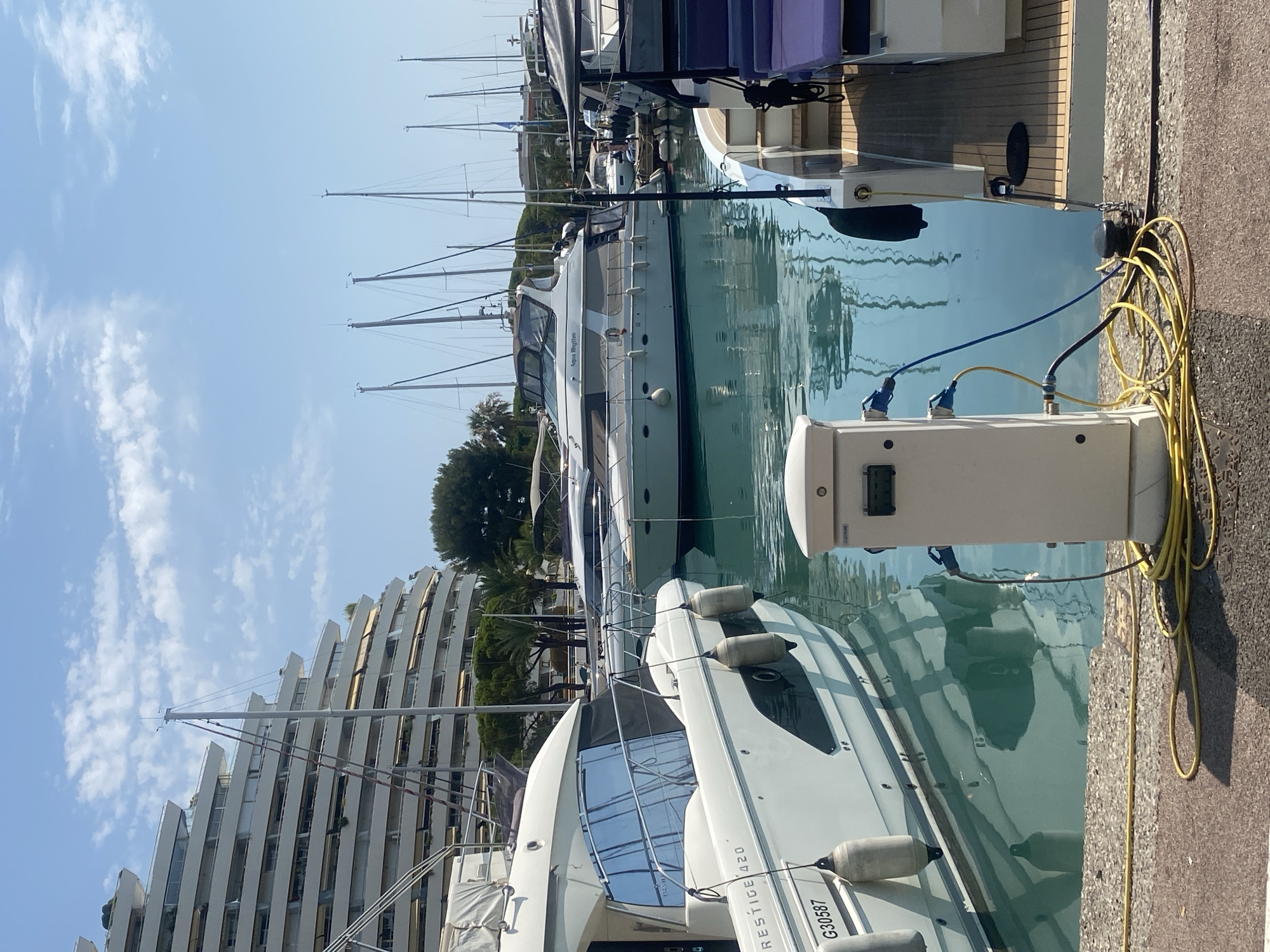 The image depicts a marina in Villeneuve-Loubet, France, under a bright, partly cloudy sky. In the foreground, a white electrical charging station for boats is visible on a paved walkway. Yellow and blue electrical cables are connected to the station and extend towards the water. To the left, a white yacht named "PRESTIGE 420" with the registration "G30587" is docked. Its reflection is visible in the calm, turquoise water. Further back, other yachts and sailboats are moored. Dominating the background is a large, tiered white building with multiple balconies, characteristic of the architecture found in coastal resorts. Lush green trees and palm trees are scattered throughout the scene. The overall atmosphere is one of a sunny day in a well-maintained harbor.
