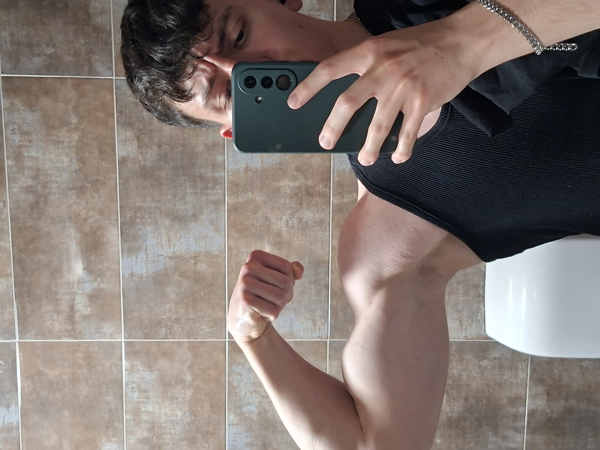A man is taking a selfie in a bathroom with tiled walls, a black tank top, and a silver chain bracelet. He is flexing his right bicep. The background includes the bathroom's beige and brown tiled walls and a white toilet. The photograph does not provide sufficient information to ascertain the precise location in Plovdiv, Bulgaria.
