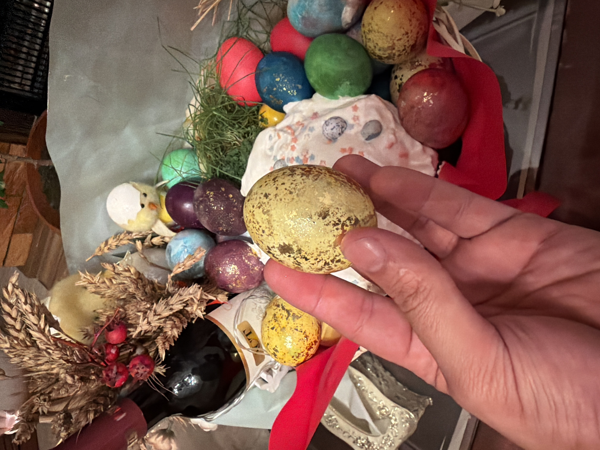 A close-up captures a hand holding a gold-speckled egg in front of a basket filled with similarly decorated Easter eggs. The basket contains red, blue, green, and purple eggs, along with a small white chick figurine and grassy elements. A red ribbon and dried wheat are also visible in the basket. A bottle with a dark liquid is present within the basket's arrangement. The environment suggests an indoor setting. There is no indication of the location being Krasnoyarsk, Russia.