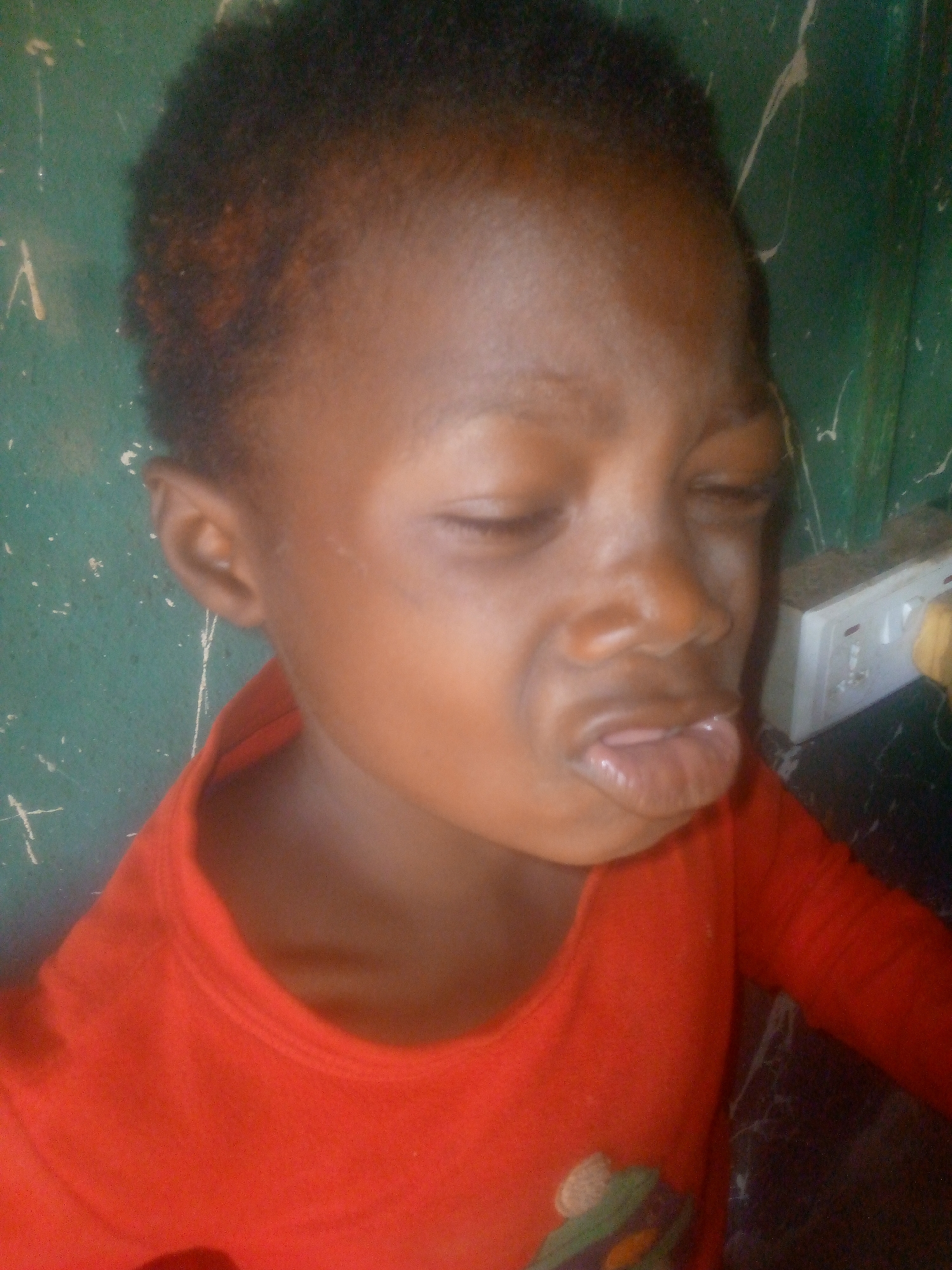 A close-up, slightly off-center shot features a young child with dark skin and short, dark, curly hair that has visible reddish-brown tones. The child is wearing a red, long-sleeved shirt with a ribbed collar.
The child's eyes are tightly closed or squinting, and their mouth is open, with lips pursed and contorted into an expression that suggests crying, discomfort, or a strong sensory reaction, such as tasting something sour. The tongue is partially visible inside the mouth.
The background is a textured, greenish wall, possibly made of painted concrete or wood, with numerous white streaks and marks. To the right of the child, a white electrical outlet, featuring two switches and two sockets, is mounted on the wall. The setting appears to be indoors, under ambient lighting. There is no discernible text in the image.