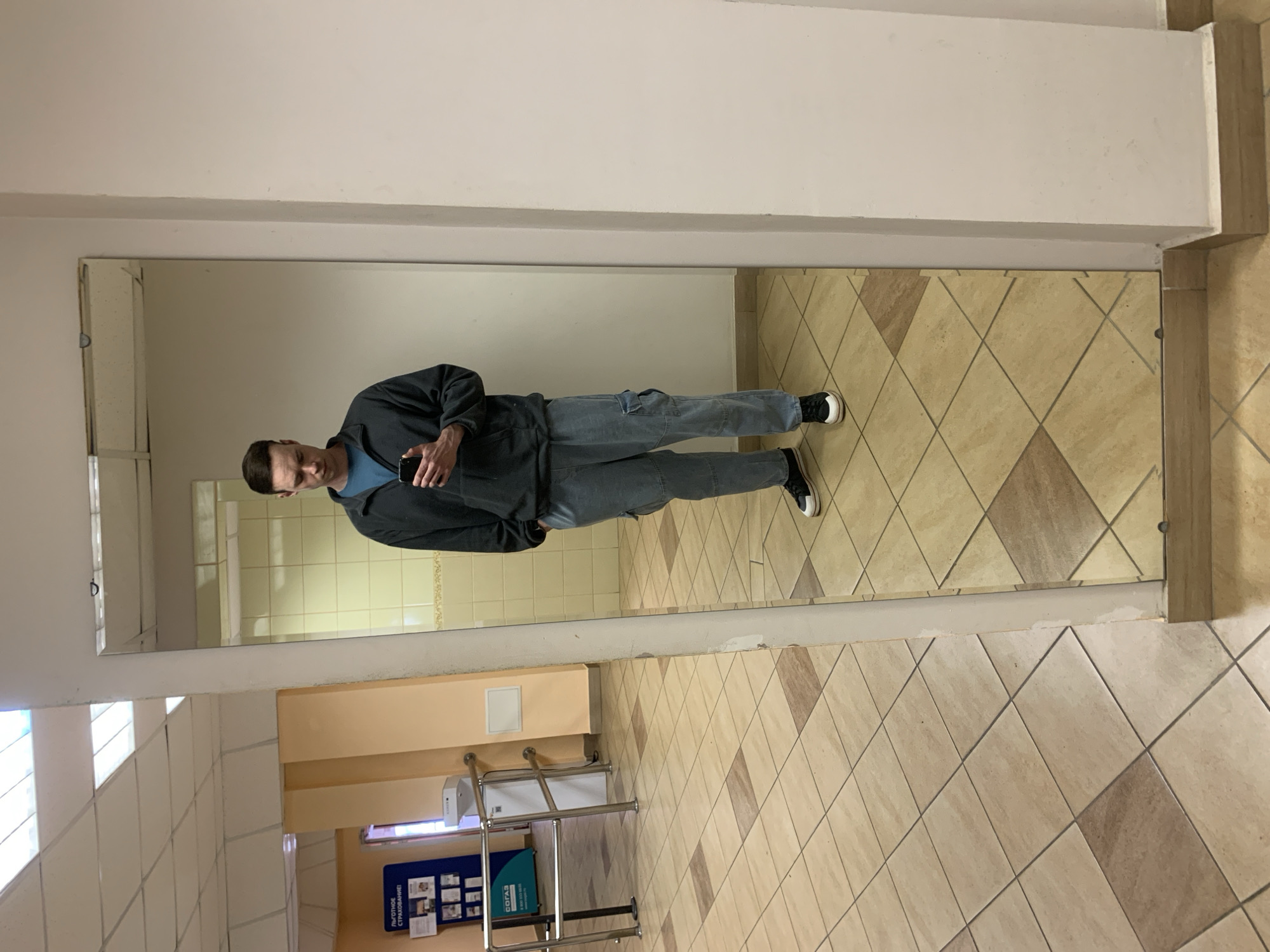 A man stands in front of a large mirror, seemingly taking a photo of himself. He's wearing a blue shirt, a black jacket, and light-colored cargo pants. He is also wearing glasses. The background reveals a public restroom with beige tiled walls and floors. In the reflection, there are bathroom stalls with a tiled wall. In the corner of the mirror is a metal handrail, next to a display with posters in Russian. The location is likely in a public building in Chaikovsky, Russia. The man is holding a smartphone.