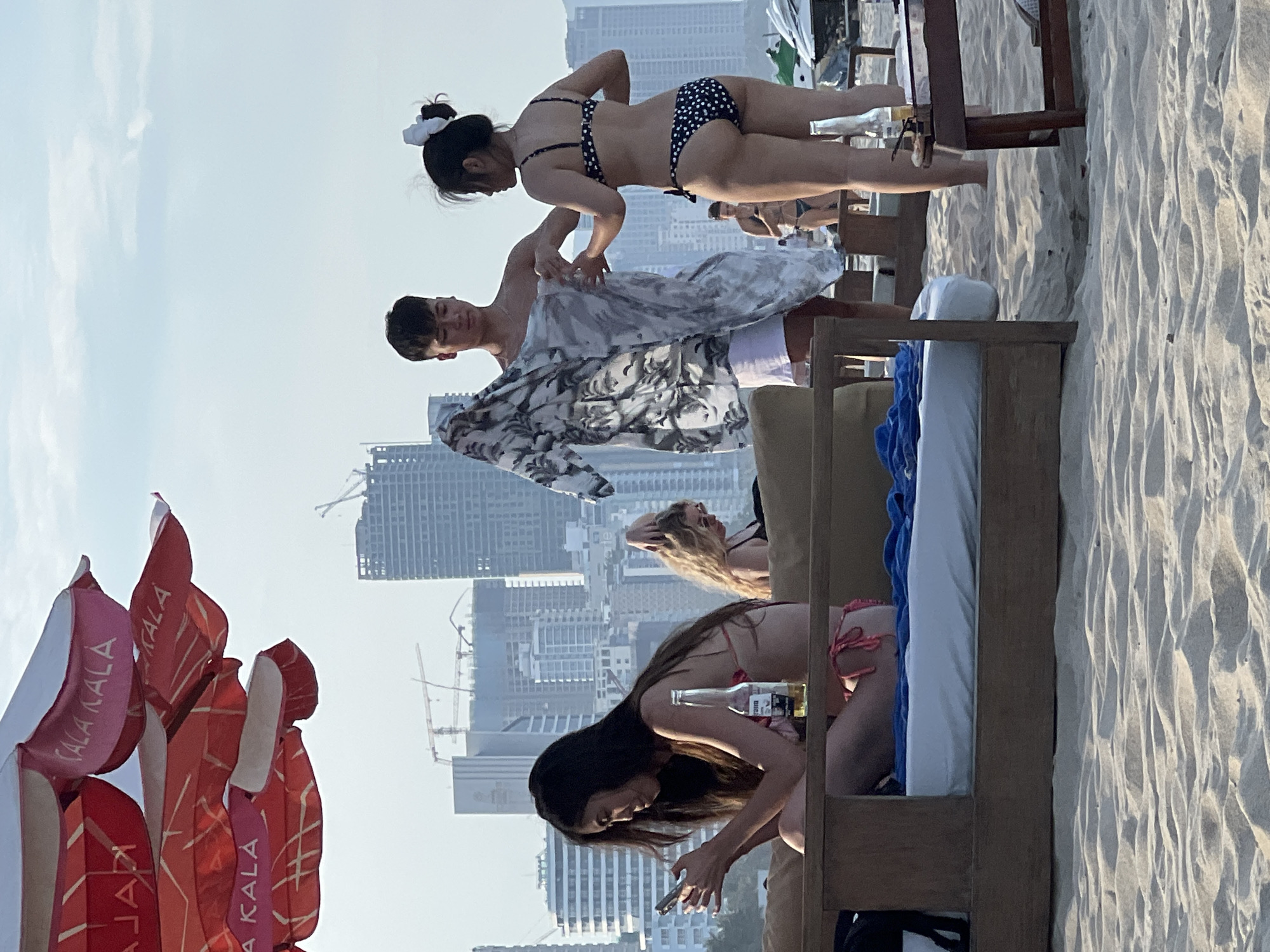 The image depicts a beach scene in Da Nang, Vietnam, with a backdrop of modern skyscrapers under construction. In the foreground, a woman in a pink bikini is seated on a wooden lounge chair, engrossed in her smartphone. To her right, a bottle of Corona beer sits on the chair. Behind them, a man and a woman in bikinis are interacting. The man is holding a patterned fabric, possibly a sarong or beach towel, as if to offer it to the woman, who is looking towards him. Further in the background, another woman in a bikini is visible. The beach is covered in fine sand, and a large, branded umbrella with "Cala Kala" text is partially visible in the upper left corner. The sky is overcast, suggesting it might be late afternoon or early evening.