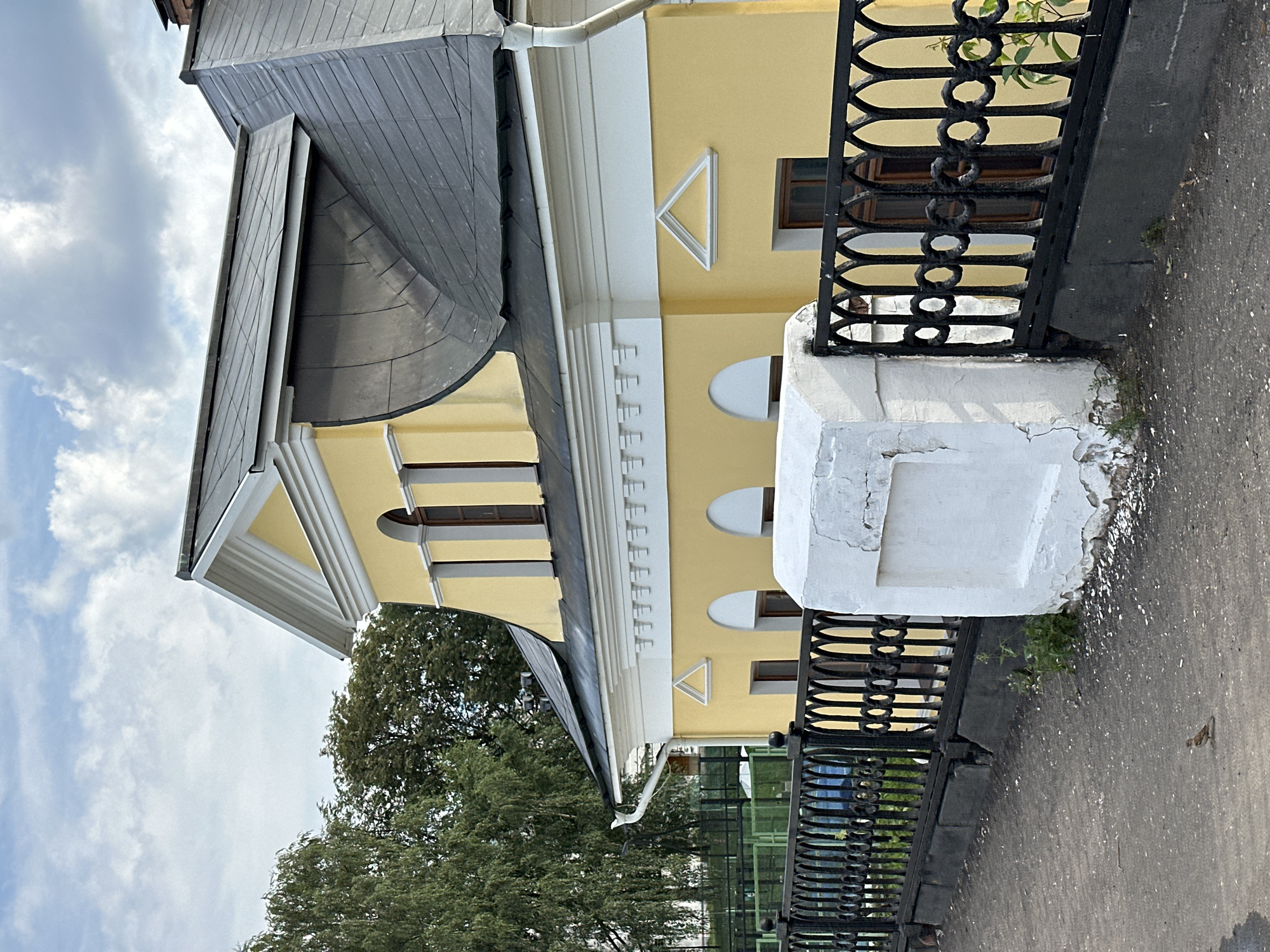 A yellow building with a dark grey roof is partially visible from the side, with trees and a cloudy sky in the background. The building has ornate white trim under the roofline, arched windows, and triangular architectural details above some windows. A black metal fence with decorative circular elements runs in front of the building. A white concrete pillar with a cracked surface stands next to the fence. The ground in front of the fence is paved with asphalt. A small bird is standing on the pavement. The location is the city of Zhukovo, Russia.