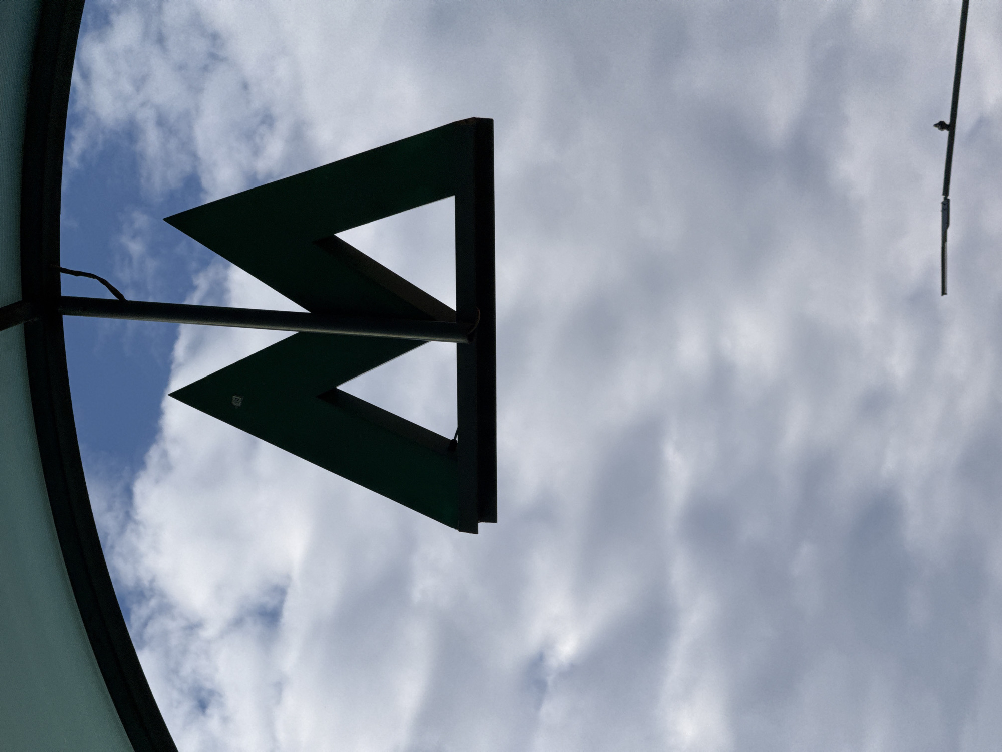 The image shows a dark green sign with the letter "M" in a stylized font, set against a cloudy blue sky. The sign is mounted on a dark pole, and a curved structure, possibly part of a canopy or awning, is visible at the top of the frame. The "M" sign is characteristic of metro station entrances in Kyiv, Ukraine. The lighting suggests it is daytime. A street lamp with a small bird perched on it is visible in the lower right corner.