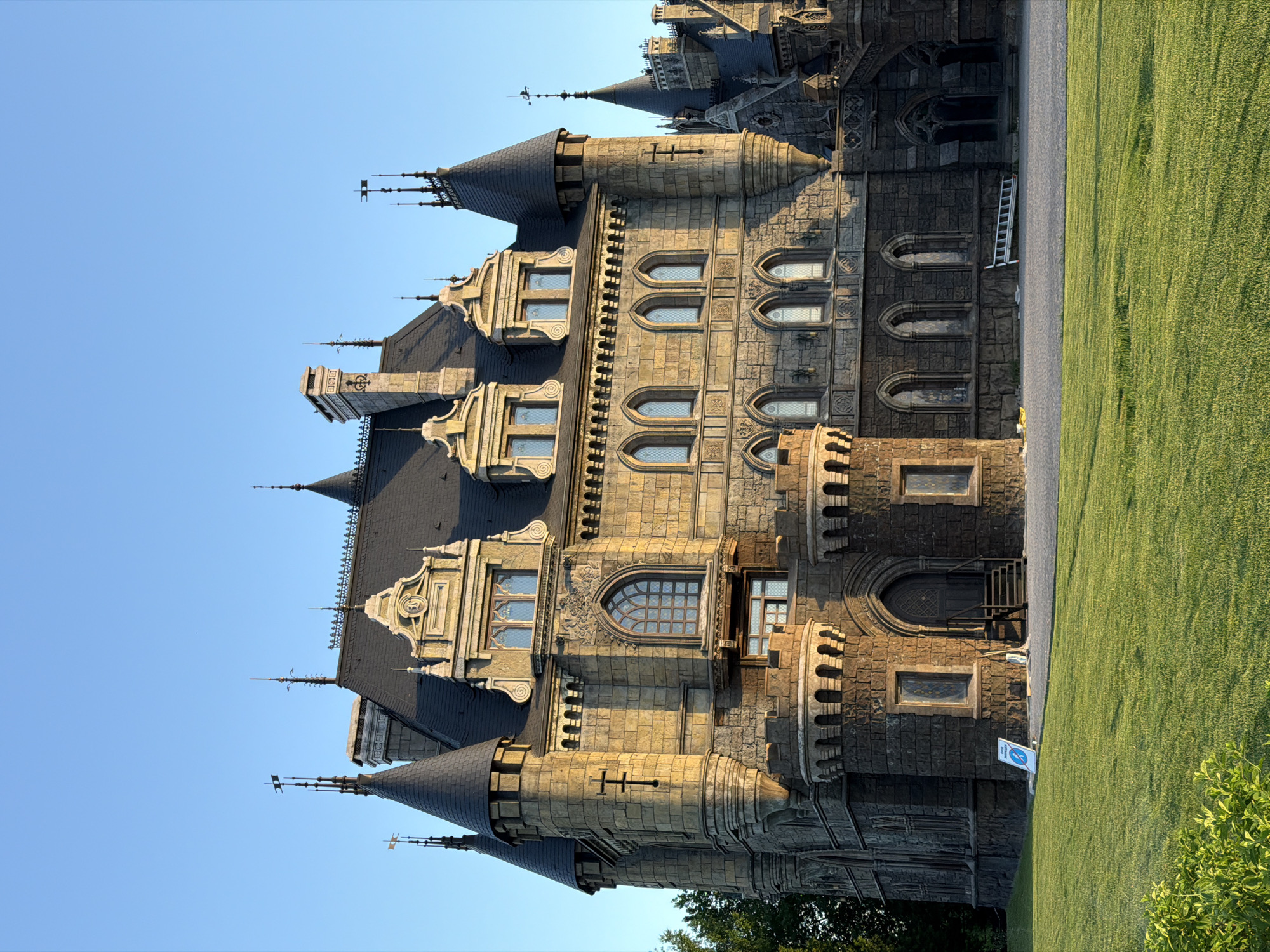 The image depicts Garibaldi Castle, a Gothic-style castle located in Khryashchevka, Russia. The castle is constructed of stone and features multiple towers, spires, and ornate windows. The sky is clear and blue. Lush green grass is visible on the right side of the frame. A ladder is propped against the building.