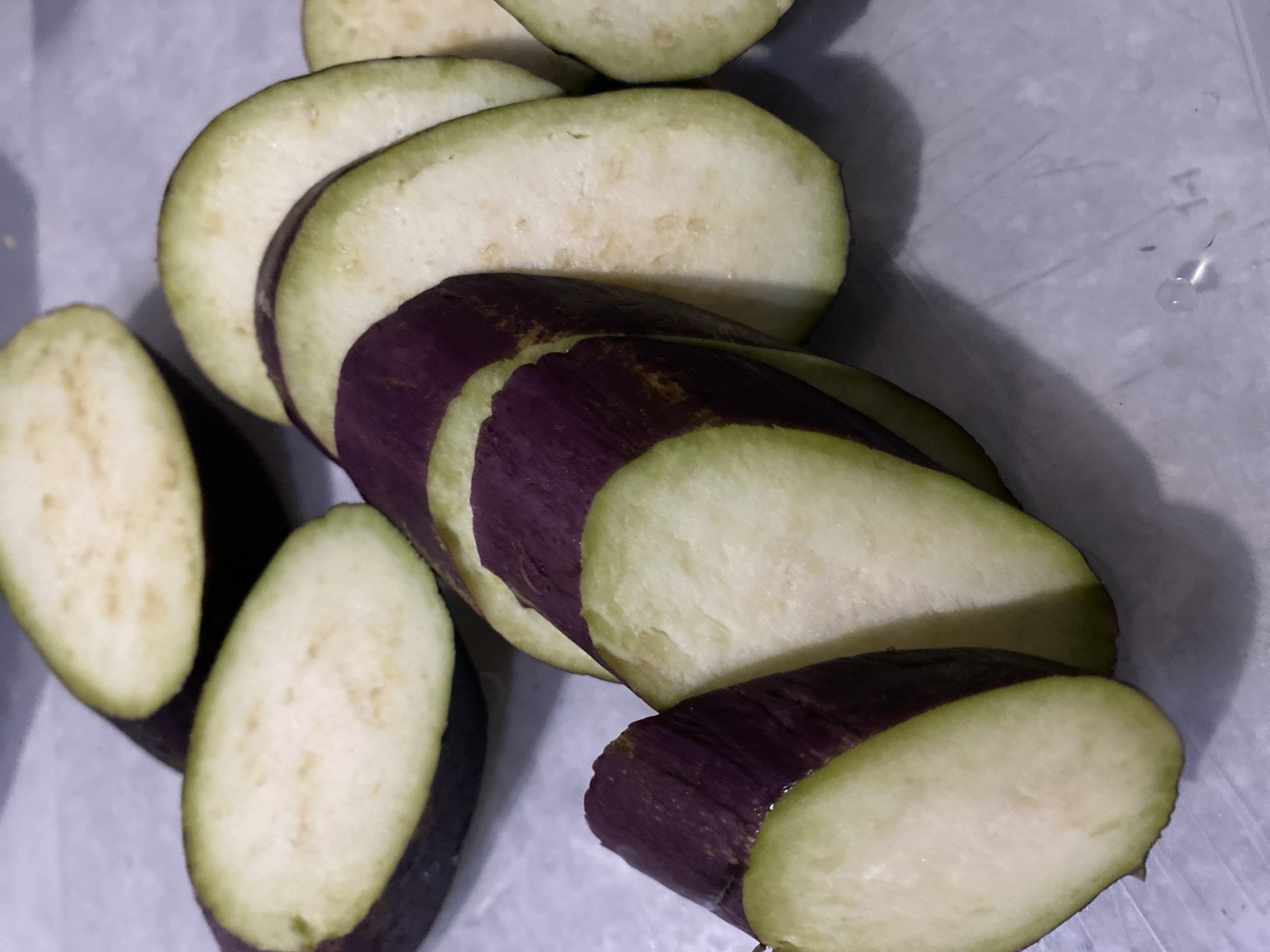 The image shows sliced eggplants on a light gray, textured surface. The eggplants are cut into roughly half-inch thick rounds, with the skin being dark purple and the flesh light greenish-white. Some slices are whole, while others are shown partially cut. The lighting is overhead, casting subtle shadows on the surface. There are no people or other objects visible in the image. The image does not contain any information about its location in Madiun, Indonesia.