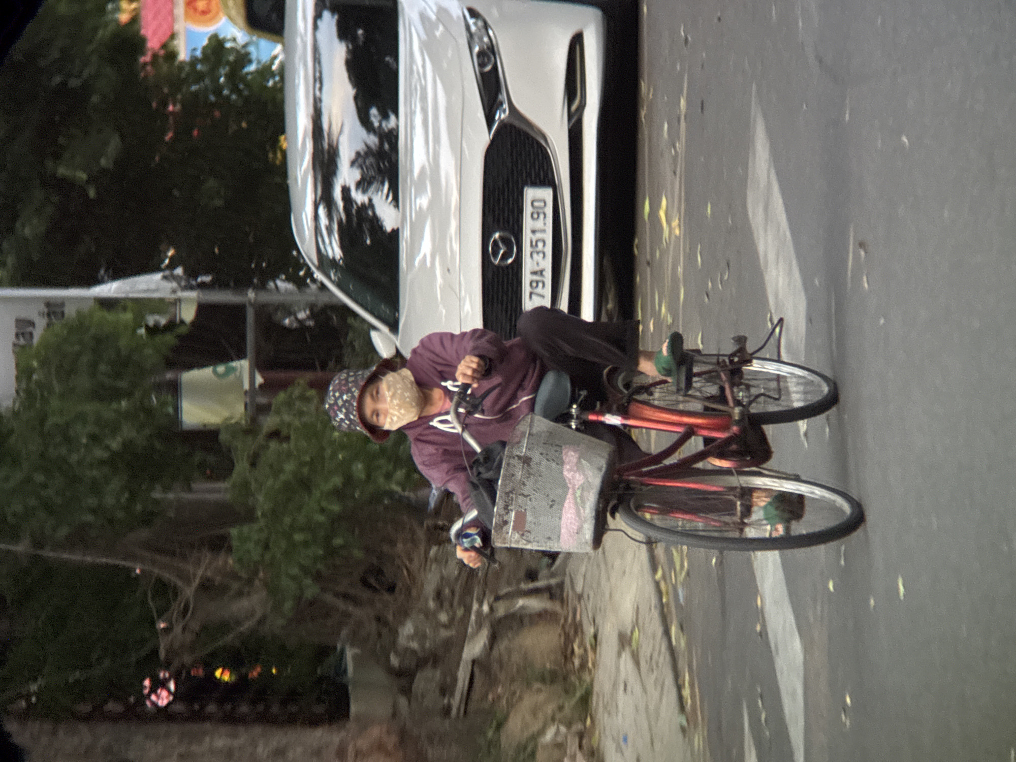 A person wearing a patterned bucket hat and a light-colored face mask rides a red bicycle with a metal basket on the handlebars down a street in Nha Trang, Vietnam. The person is wearing a purple hooded jacket and dark pants, with green sandals on their feet. The bicycle has a basket containing what appears to be a pink cloth item. Behind the bicycle, a white Mazda car with a license plate reading "79A-351.90" is visible. The street has a crosswalk marking. Trees and some signage are visible in the background, suggesting an urban environment.