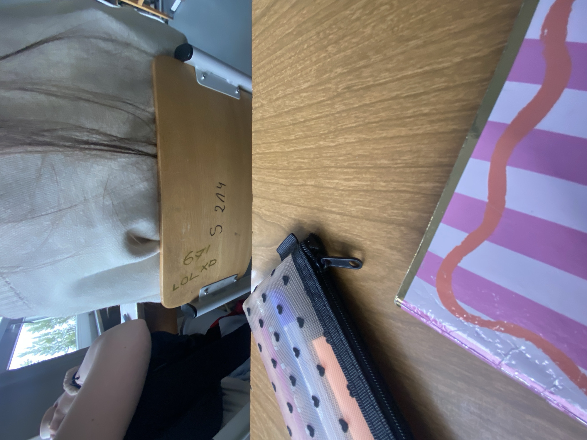 The image shows a desk with a notebook, a pencil case, and a wooden table attachment. Writing is visible on the wooden table, including "671 LOL XD" and "S. 204". Part of a person is visible in the bottom left corner. The surroundings are indeterminate, but there's a hint of a window and foliage outside. The location in the city of Unknown, Unknown is indeterminate.